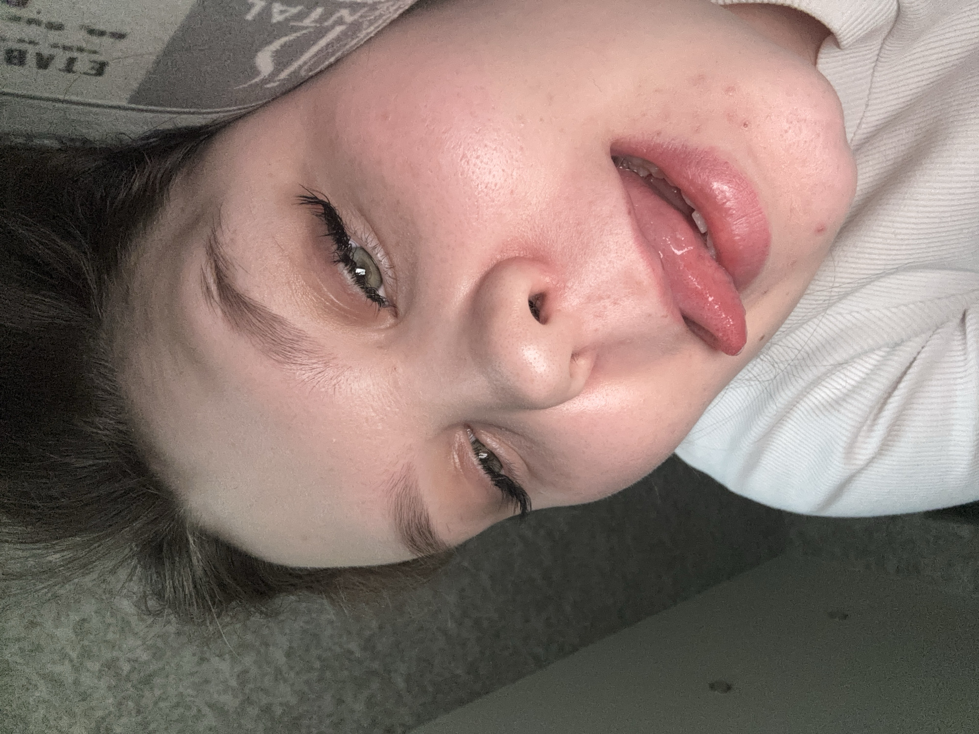 A close-up, rotated image depicts a person lying down, sticking out their tongue. The person has fair skin, light green eyes, and dark hair. The person is wearing a light-colored shirt. A gray surface, possibly a pillow or upholstered furniture, is visible in the background. The image does not provide sufficient information to determine a location in Perm, Russia.