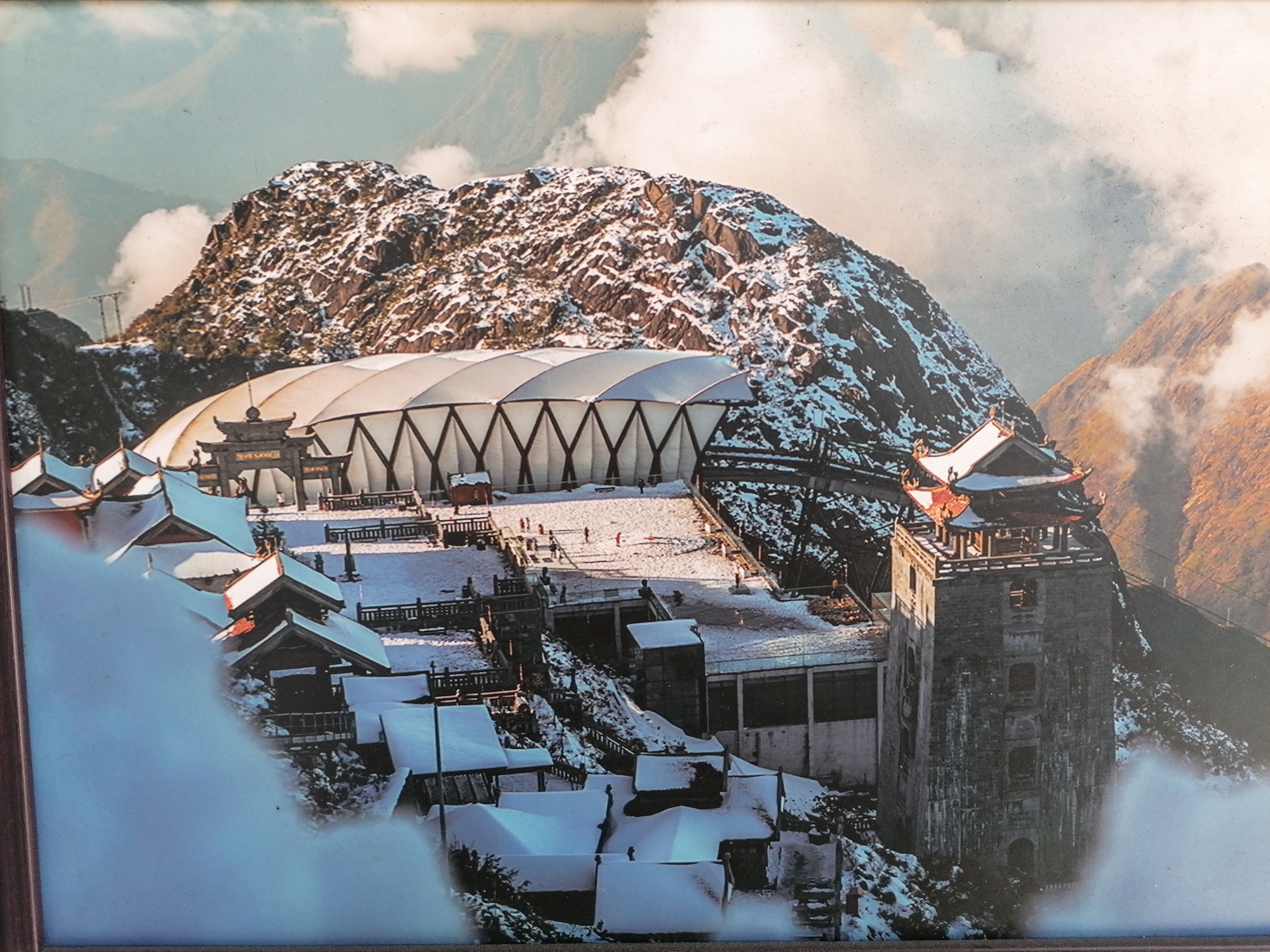 The image shows a snow-covered mountainous landscape in Sa Pa Ward, Vietnam. A large mountain dominates the background. A cluster of traditional Vietnamese-style buildings with red and white roofs, covered in snow, are in the foreground. A tall, gray, square tower stands prominently among the buildings. A modern white structure with a curved roof is positioned behind the traditional buildings. Several people are visible on a snow-covered plaza between the modern structure and the buildings. Clouds surround the scene.