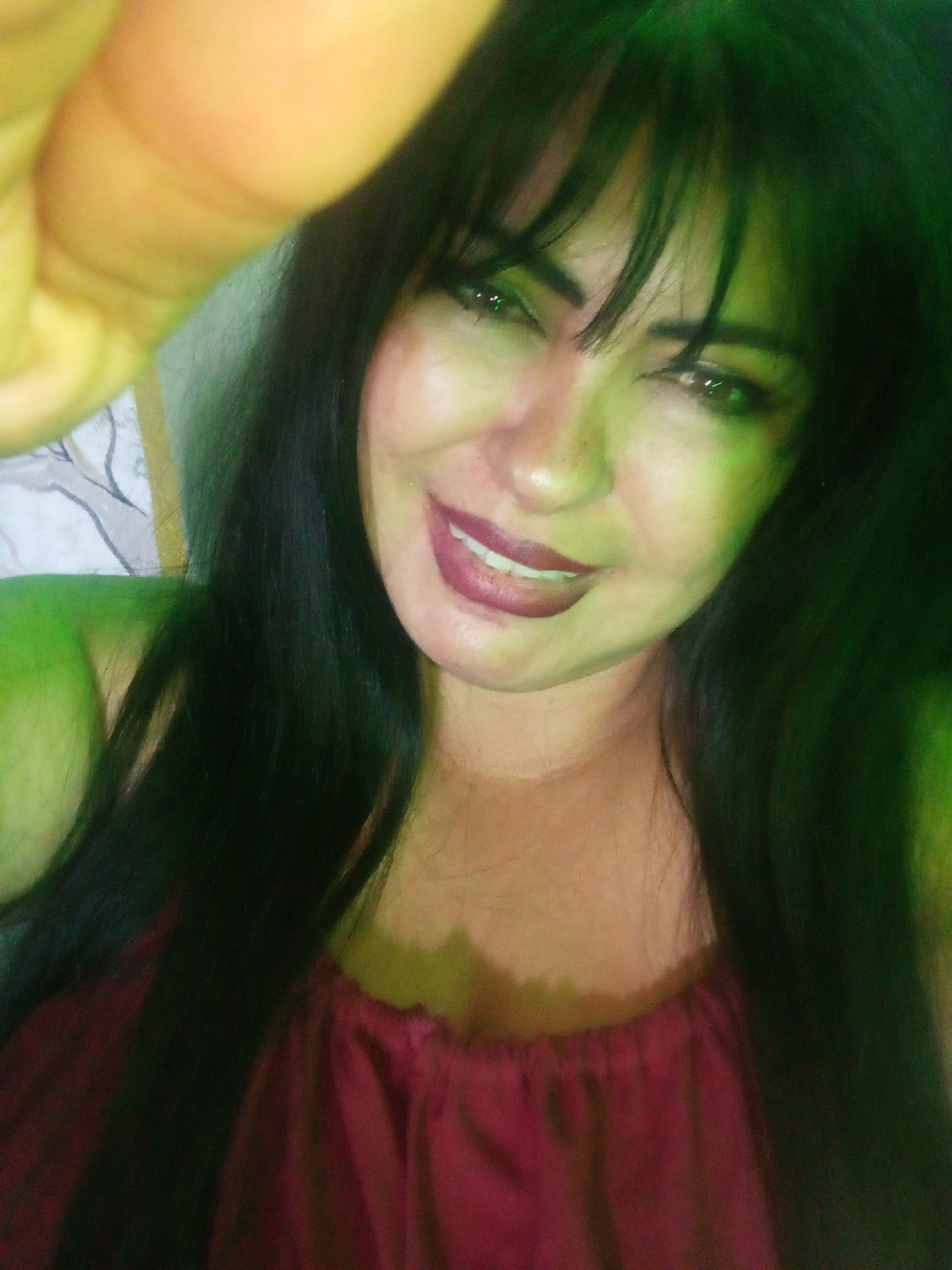 A woman with dark hair and a red top is posing for a selfie. Her left hand is partially obscuring the lens. The lighting appears to be green-tinted. The location is Unknown, Unknown.