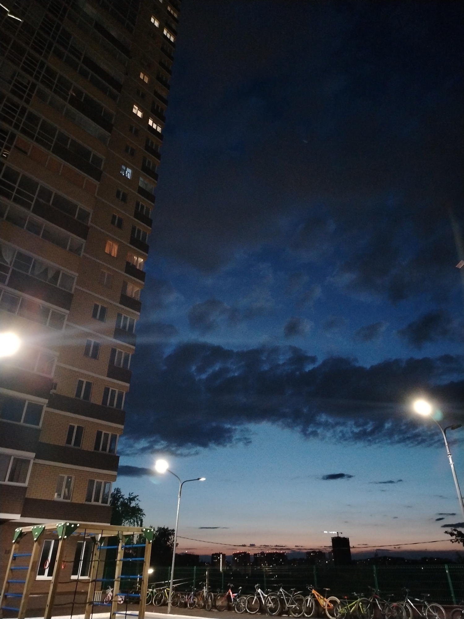 The image depicts a twilight scene, likely at dusk, in a residential area of Izhevsk, Russia. A modern, multi-story apartment building with a light-colored facade and dark balcony elements occupies the left side of the frame, its many windows showing warm interior lights from occupied units.
The sky, filling a significant portion of the image, displays a dramatic transition from deep blue overhead to lighter blues, subtle oranges, and pinks near the horizon. It is heavily overcast with textured, dark clouds, contributing to a moody atmosphere.
Two bright streetlights with white-yellow illumination are prominent, one on the lower-left near the building and another further to the right. Both generate noticeable lens flare. In the bottom left, a section of a wooden playground structure with blue metal bars and green brackets is visible.
A row of various bicycles is parked along a fence in the middle ground. Beyond this, a distant cityscape is silhouetted against the lighter horizon, featuring several buildings and faint lights, with one taller, darker building standing out. Some sparse trees and bushes are also present in the mid-distance.
The scene conveys a quiet, serene urban evening in a contemporary residential neighborhood. No specific activities or people are visible, emphasizing the stillness of the moment. No discernible text appears in the image.