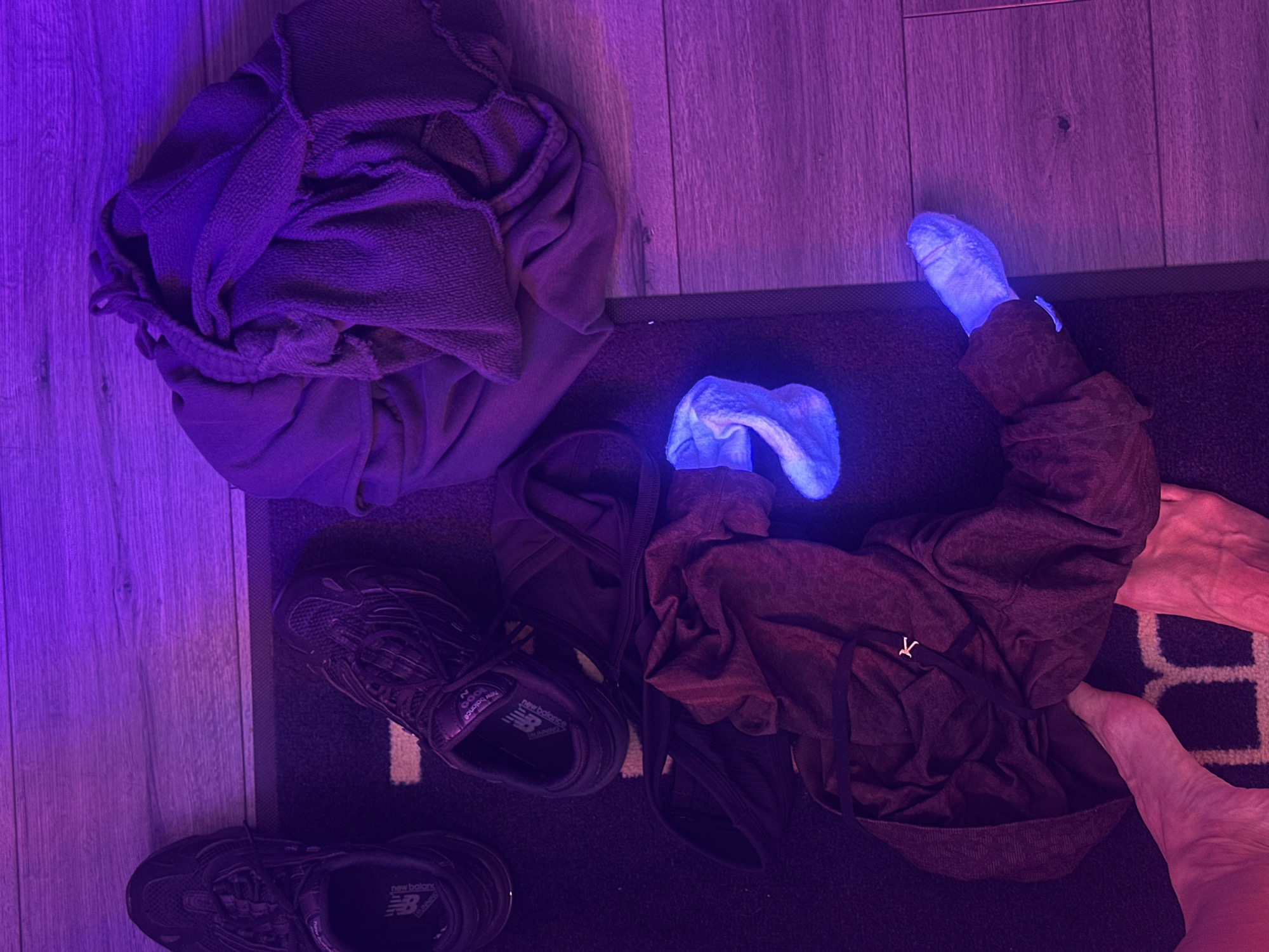 The scene depicts a collection of items on the floor. A pile of folded or crumpled gray clothing is visible on a wood-grain surface, beside a dark rug. On the rug are a pair of black New Balance running shoes, a dark-colored hoodie, and two socks. One sock appears crumpled and partially tucked into the hoodie. A person's bare feet are near the edge of the rug. The entire scene is bathed in purple/blue light. There is no indication of Tunbridge Wells, United Kingdom.