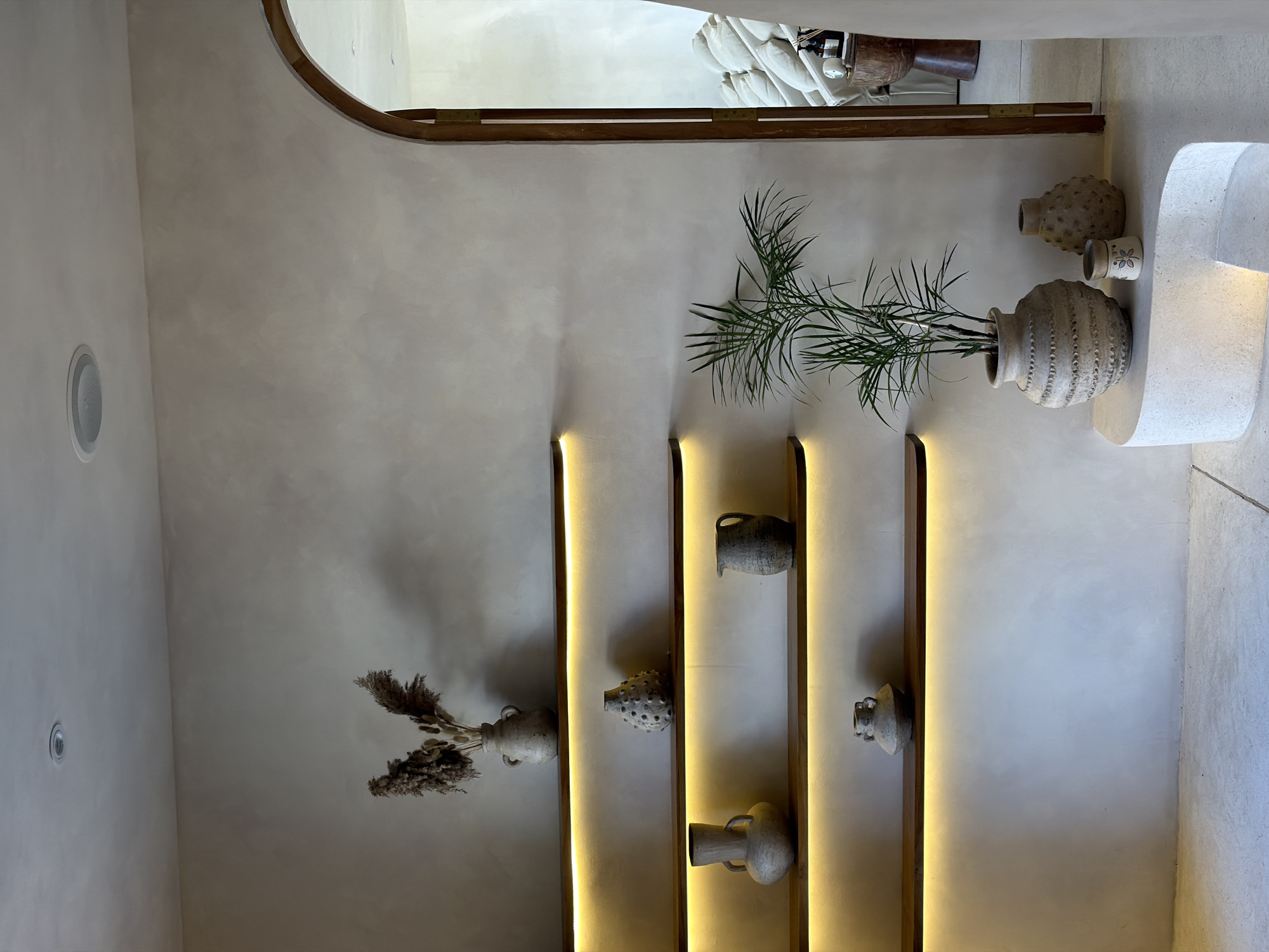 The image depicts an indoor scene, likely a home or a commercial space, in North Kuta, Indonesia. The focus is on a wall with several illuminated shelves displaying various decorative ceramic vases and dried floral arrangements. To the right, an arched doorway leads to another room where a neutral-colored sofa is visible. In the foreground, a potted palm plant stands next to several textured ceramic vases on a white, organically shaped pedestal. The overall aesthetic is minimalist and earthy, with a neutral color palette dominated by beige and off-white tones, complemented by warm, soft lighting. No people are present in the scene.