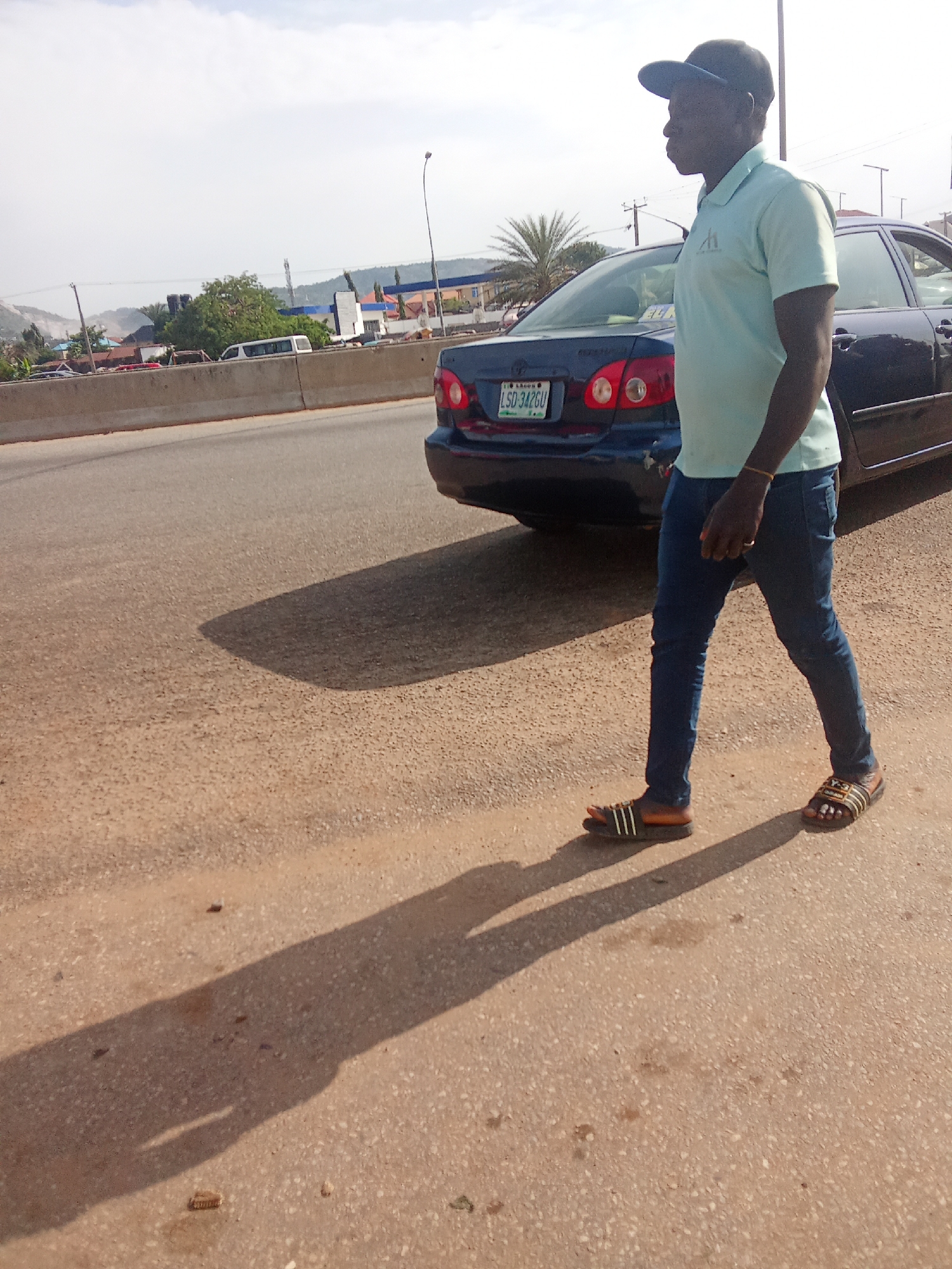 A side view of a dark blue sedan is shown parked on the side of a road in Kagini, Nigeria. A man wearing a light blue polo shirt, dark jeans, a blue baseball cap, and sandals walks away from the camera in front of the car. The car's license plate reads "LSD 342GU". In the background, buildings and trees are visible, along with a cloudy sky. The man's shadow is cast on the asphalt road.