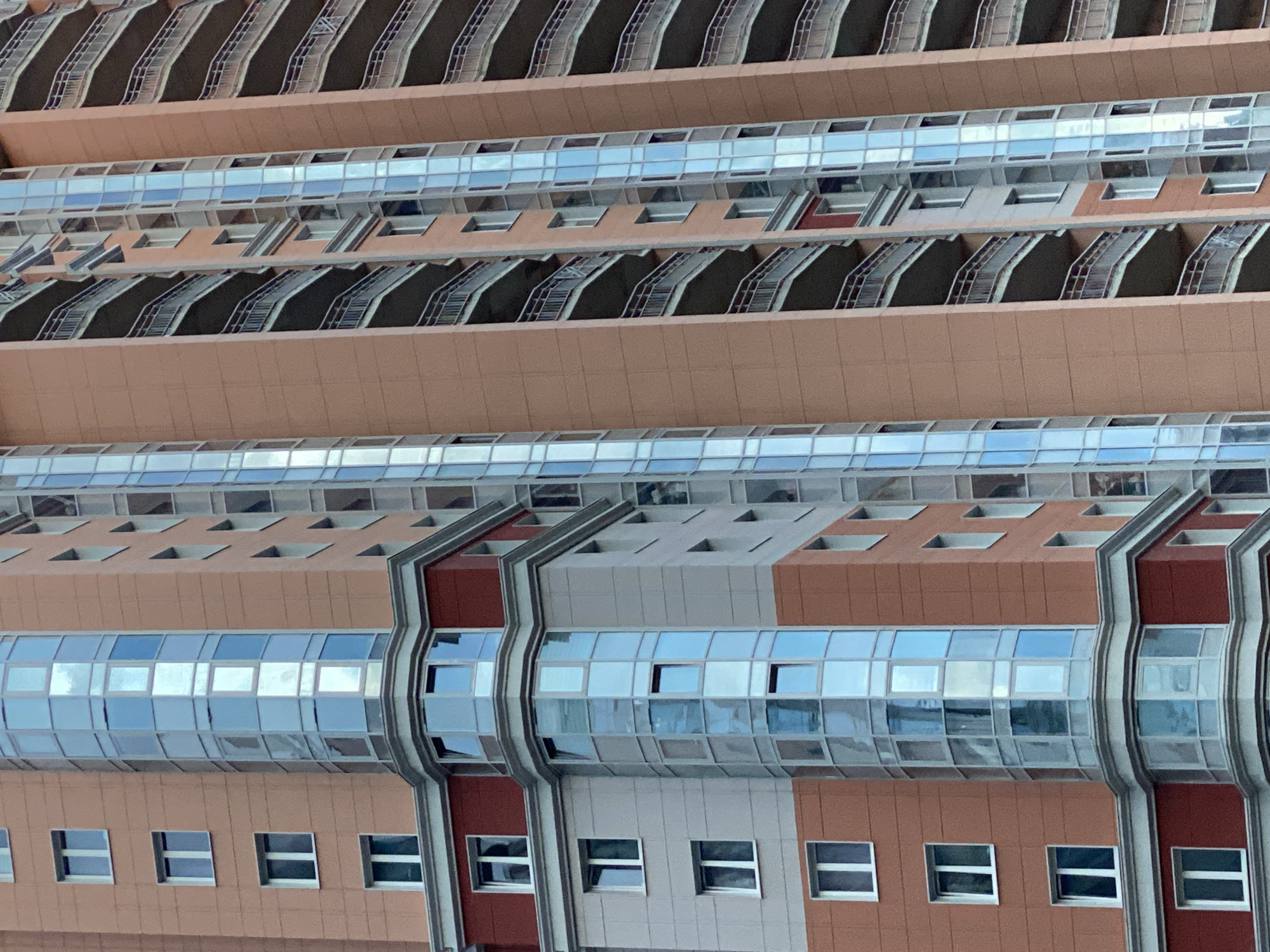This is a close-up, medium-shot, eye-level, outdoor, daytime, vertical, full-frame, eye-level, candid photograph of a section of a modern, tall apartment building in an urban environment. The building's exterior is predominantly a salmon or peachy-orange color, with sections of grey and deep red. The architecture features multiple floors, each with numerous windows. Many of these windows are part of vertical, multi-story glass columns that extend upwards, reflecting the sky and surrounding environment with a bluish tint. These glass sections are segmented into smaller panes, creating a patterned effect.
Interspersed between the glass columns are balconies with metal railings, visible on several floors. These balconies appear to be recessed into the building's facade. There are also rectangular windows that are set directly into the colored walls of the building, these are white-framed. The building has a complex, angular design, particularly evident in a corner section with a bay window or a protruding glass structure. White decorative trim lines some of the architectural divisions between colors and levels. No people are visible in the frame. The overall impression is one of contemporary urban residential architecture. The location is identified as Unknown, Russia.