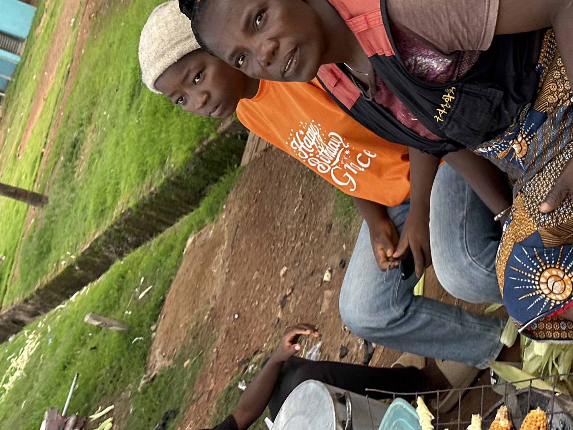 This media file shows three people in an outdoor, dusty setting with green grass visible in the background. Two individuals are in the foreground, a child and an adult, both appearing to be of African descent. The child, on the left, wears a light-colored knit cap and an orange t-shirt. The adult, to the right, wears a dark vest over a patterned fabric and blue jeans. Their faces are partially visible, with the adult looking towards the camera. A third person, likely an adult, is partially visible on the right, wearing a patterned garment. The scene appears to be a casual gathering or marketplace, possibly in Jos, Nigeria, given the location context.
The people seem to be engaged in some form of activity, though it is not explicitly clear. The child's t-shirt has white lettering that reads "Happy Birthday Grace". The adult on the right has their hands visible, holding something that could be related to the activity, and is wearing a bracelet. The overall mood appears to be relaxed. The lighting suggests it could be daytime, possibly late morning or afternoon. The ground is a mix of dirt and some debris. In the lower right corner, there are some ears of corn and a metal grill or rack, hinting at food preparation or sale. The overall impression is of a candid moment captured in a local environment.