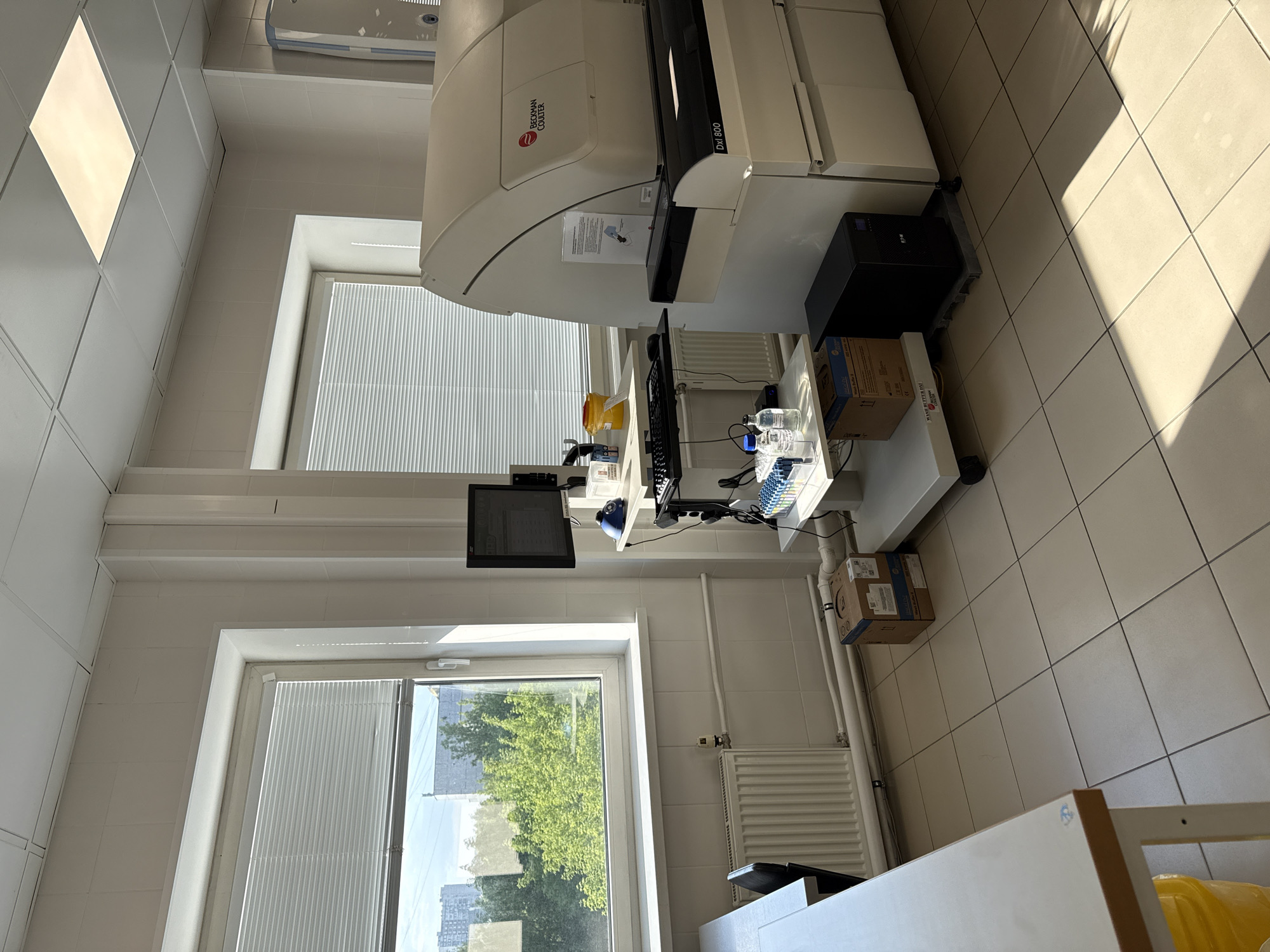 The image shows the interior of a laboratory in Moscow, Russia. A Beckman Coulter DxI 800 immunoassay analyzer dominates the scene, positioned near a window with white blinds. A computer monitor and keyboard are visible on a small table adjacent to the analyzer. Two cardboard boxes are on the floor beneath the table. A second window is visible in the background, revealing a view of trees and buildings, typical of a residential area in Moscow. The room has white tiled walls and floors, illuminated by overhead fluorescent lights and natural light from the windows. A radiator is beneath the window. A partial view of a white countertop is visible in the lower right corner of the image, along with a yellow container.