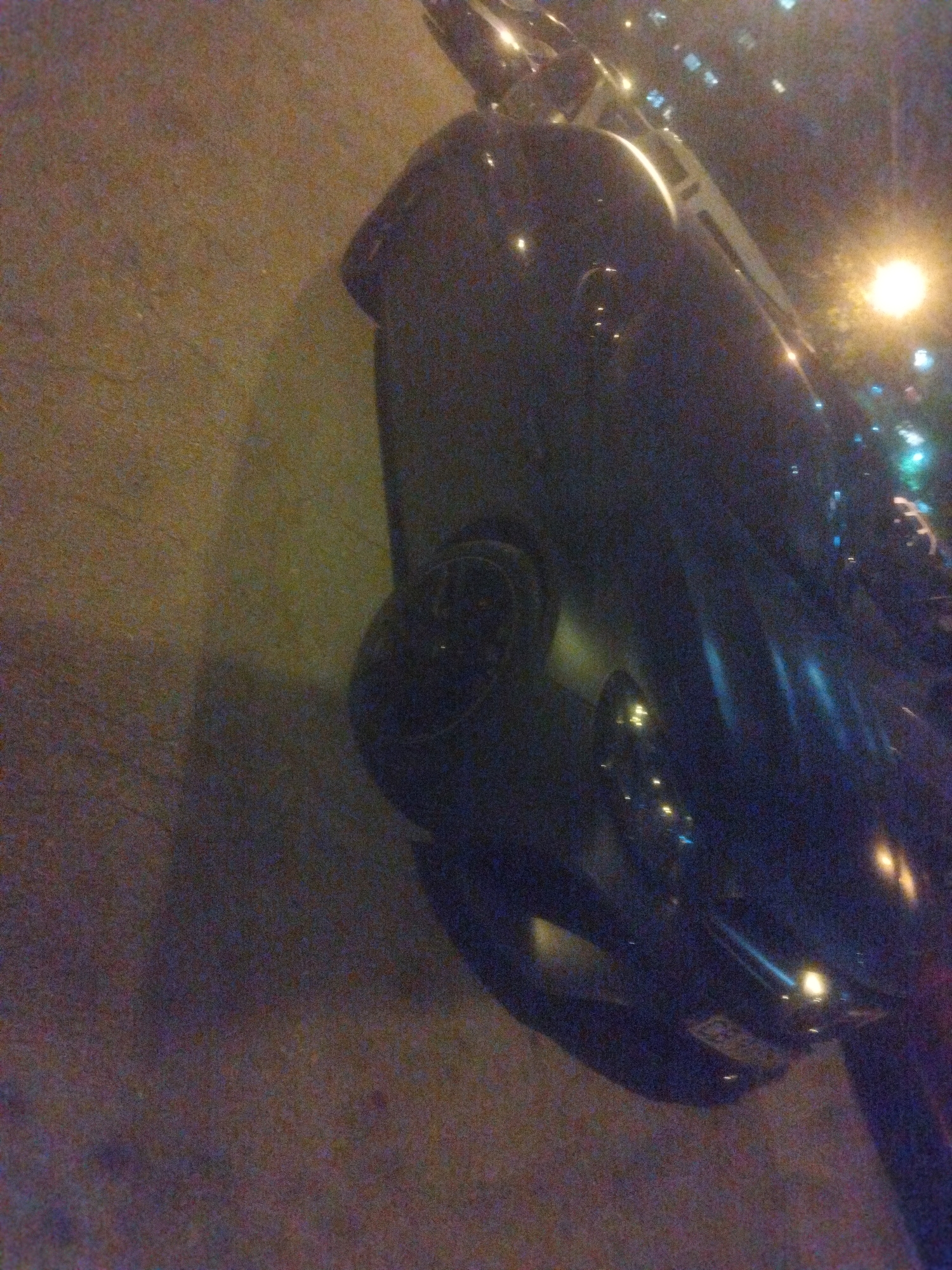 The image depicts a dark-colored car parked on a paved surface, likely a street, at night. Streetlights and other building lights are visible in the background, suggesting an urban environment. Based on details from the license plate the location is in the city of Sofia, Bulgaria. The focus is on the rear of the car, with a wheel visible and light reflecting off the body.