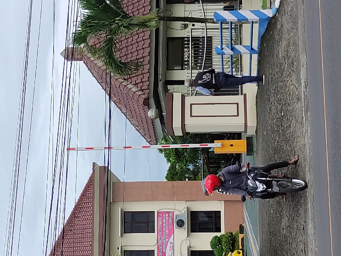 The image depicts an outdoor scene at what appears to be a guarded entrance or checkpoint in Kepanjen, Indonesia, under an overcast sky during the daytime.
In the foreground, a person wearing a grey jacket and a red helmet is seated on a black motorcycle, facing towards the right. Their left foot is on the ground, suggesting they are stopped or waiting. The motorcycle is positioned in front of a red and white striped boom gate, which is lowered horizontally across the path. The boom gate mechanism, a yellow box, is visible on the right side of the road.
Behind the boom gate and to the right, another individual stands, facing away from the viewer. This person is wearing a dark jacket with a white design on the back, dark pants, and shoes. They have light-colored hair or are wearing a light-colored head covering. They appear to be an attendant or guard.
The background features two buildings. The building behind the motorcyclist has a reddish-brown tiled roof and light brown walls with dark-paned windows. A pink sign with unreadable text, likely in Indonesian, is visible on the wall below a window. Further right, behind the standing person, another part of the building complex is visible, featuring blue and white striped railings. A green palm tree rises above the tiled roofs. Numerous black electrical or communication wires crisscross the upper portion of the frame against the cloudy sky. The ground is paved.