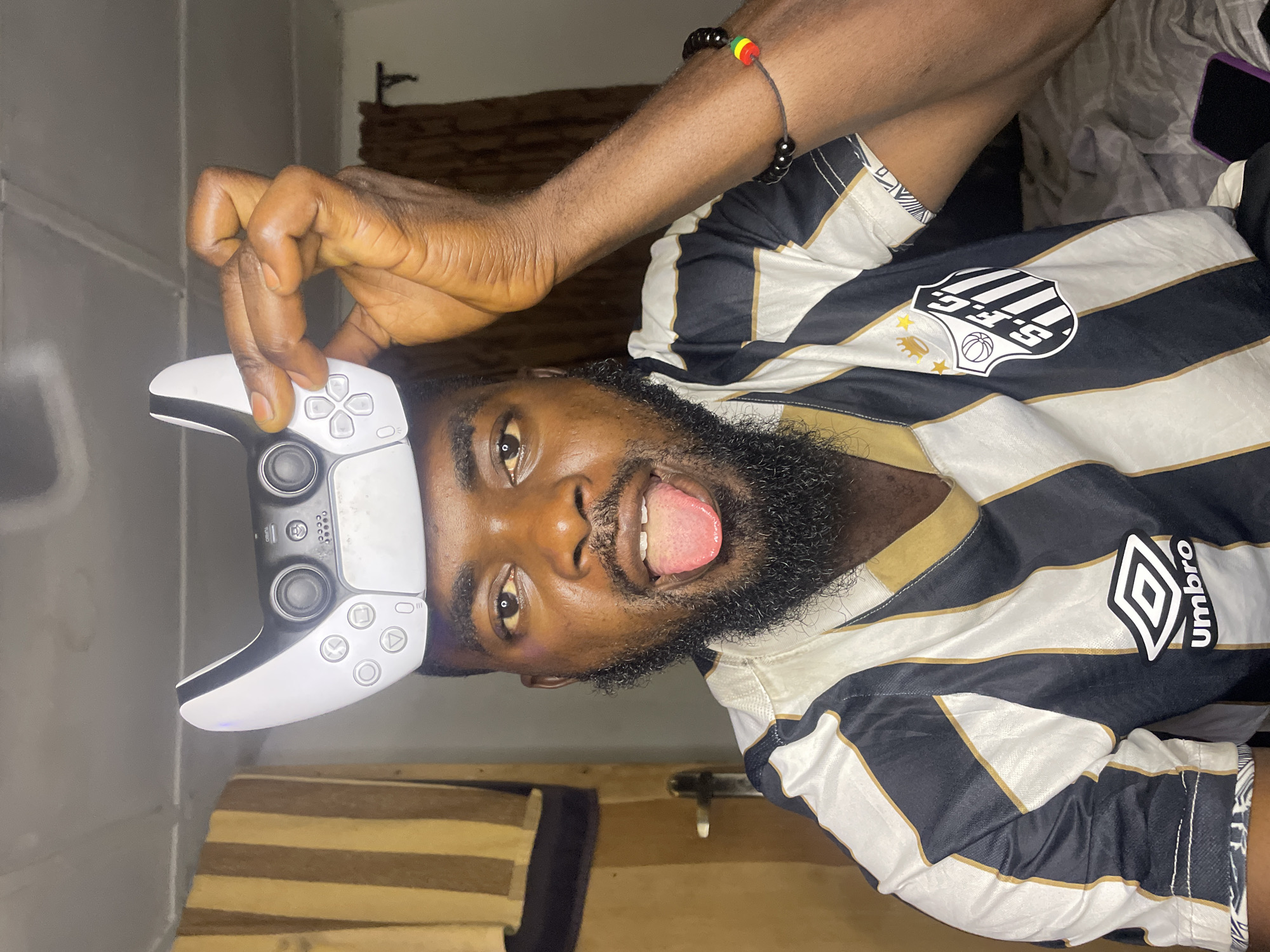 A person is holding a PlayStation 5 controller above their head with one hand. The person has dark skin, a beard, and is sticking their tongue out. They are wearing a striped black and white jersey with "S.F.C." and an Umbro logo visible. The person is wearing a bracelet that has the colors of the Rastafari flag on it. The background includes a striped curtain and a bed. This image was taken in Jos, Nigeria.