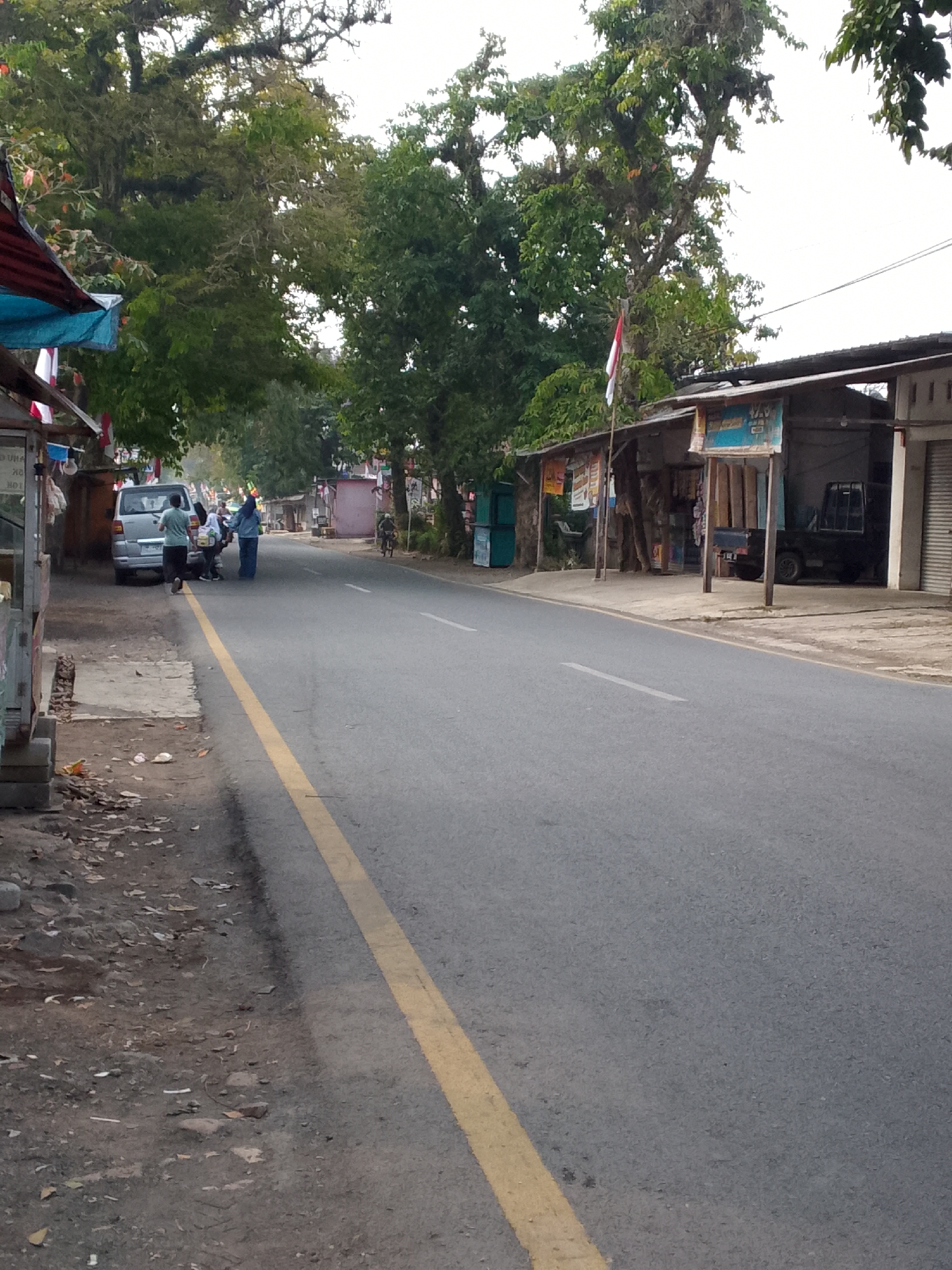 The image depicts a street scene in Haurwangi, Indonesia. A paved road runs through the frame, marked by a yellow line on the right side. On the left side of the road, there is a sidewalk area with dirt and scattered leaves. Adjacent to the sidewalk are several small stalls or shops with awnings and hanging decorations.
In the foreground on the left, a portion of a food stall or shop structure is visible, made of concrete and wood. Further down the sidewalk, a silver van is parked. Three people are standing near the van; one person, wearing a light blue shirt and dark pants, is walking away from the viewer and the van. Two other individuals, one in a blue headscarf and long blue garment, and another in a white shirt and dark pants, are facing each other and appear to be interacting.
Across the street, on the right side of the road, are more shops or buildings. One structure has a dark-colored pickup truck parked inside a covered area. Another building has a corrugated metal roof. A flagpole with an Indonesian flag is visible between two of the buildings on the right. A person on a bicycle is riding in the distance on the right side of the road. Trees with lush foliage overhang the street and buildings, creating a natural canopy. The sky is overcast.