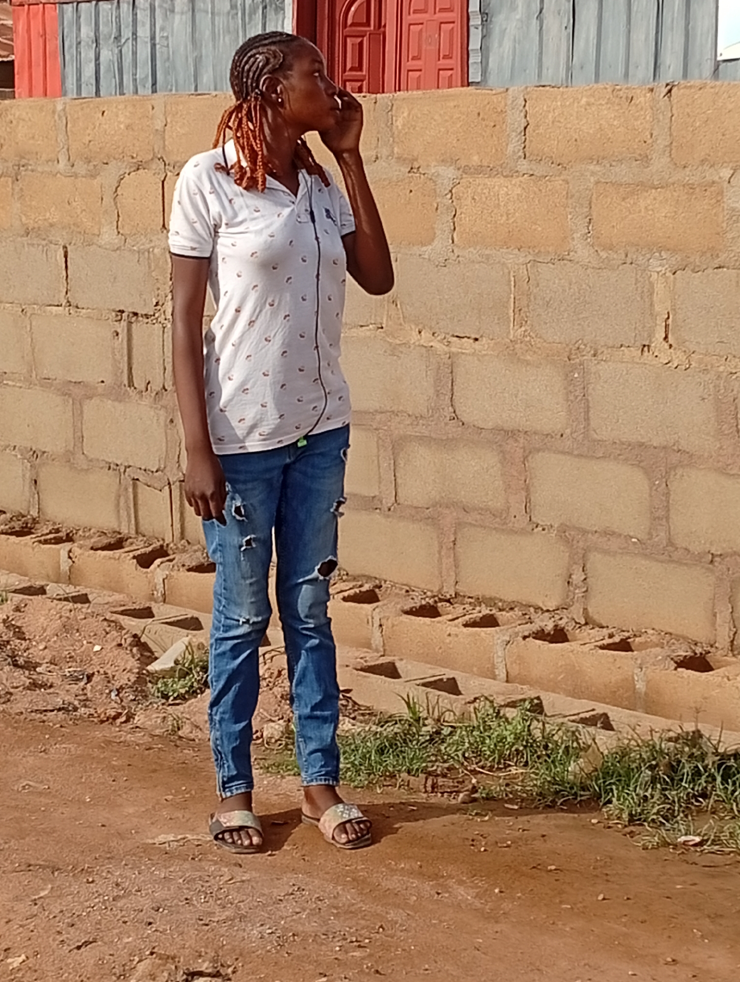 A woman stands on a dirt path in Sabon Tasha, Nigeria. She wears a white polo shirt with a pattern, distressed blue jeans, and sandals. Her hair is styled in braids with reddish-brown extensions. She is holding a phone to her ear. Behind her is a cinderblock wall with a corrugated metal fence and a red door in the background. Small patches of grass grow along the base of the wall.