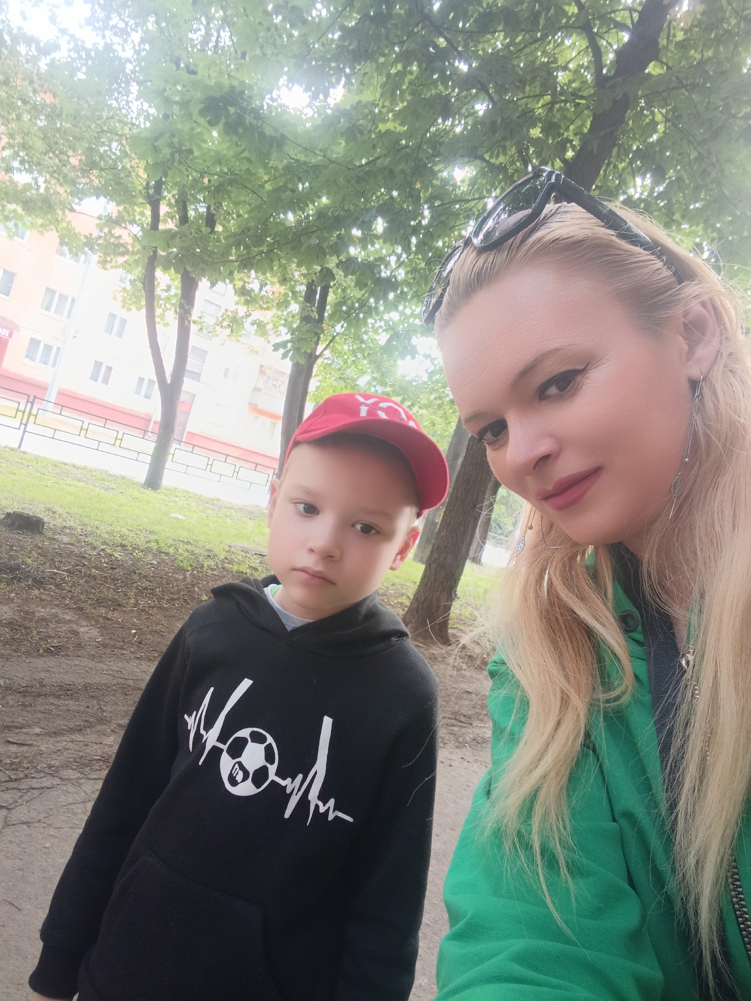 In a medium-shot, a blonde woman wearing a bright green hooded jacket and sunglasses is taking a selfie with a young boy. The boy, on the left side of the frame, is wearing a red baseball cap and a black hoodie with a white soccer-themed graphic. They are outdoors, with trees and a building visible in the background. The building appears to be a residential apartment block with multiple floors and windows. The overall setting suggests an urban park or a residential area in Chernihiv, Ukraine. The ground is paved with dirt and some debris, and green grass is visible in the background. The lighting is somewhat diffused, indicating overcast conditions or a bright day with sun filtering through trees.