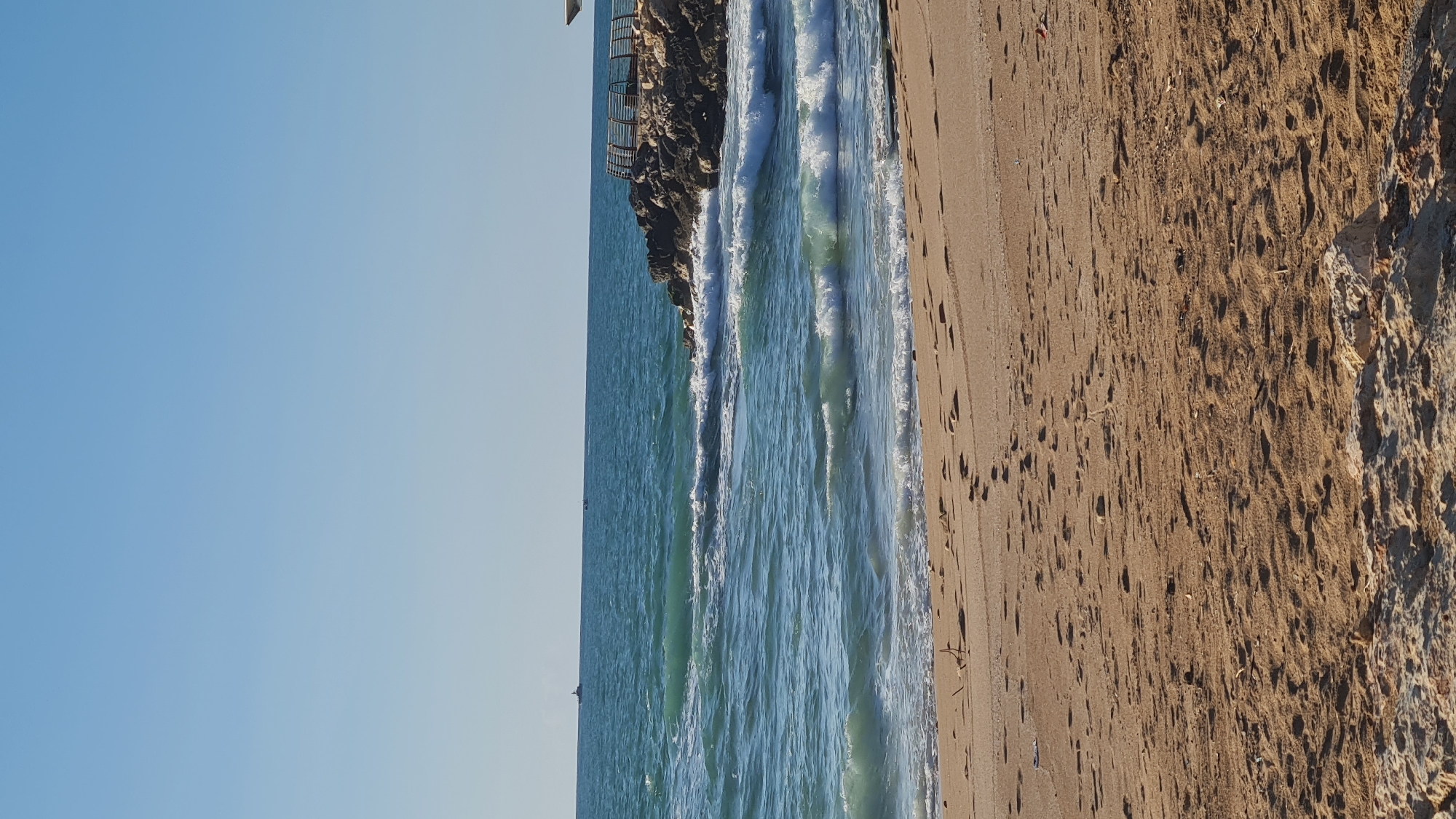 The image captures a coastal scene in Nahariyya, Israel. The sky is clear and light blue. To the right of the sky, there is a beach with sand and rocks. Ocean waves crash against the shore, next to a rocky cliff with a railing at the top.