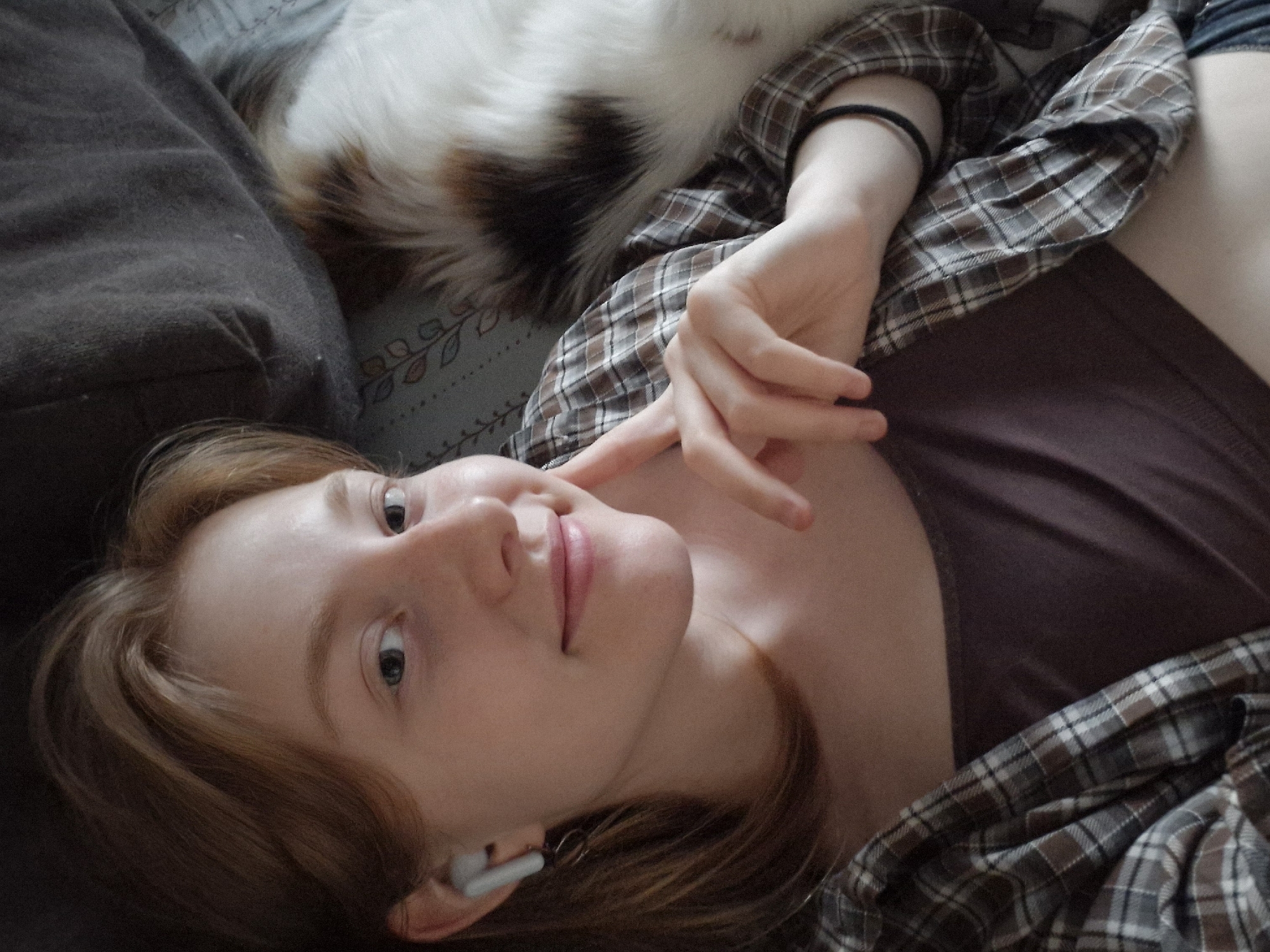A young woman lies on a bed with a brown pillow to her left and a white and brown cat above her. She wears a brown t-shirt and a plaid button-down shirt. An earbud is in her left ear, and her left hand is near her face. The bedding underneath has a floral design. The city of Yekaterinburg, Russia, is not discernible from this image.