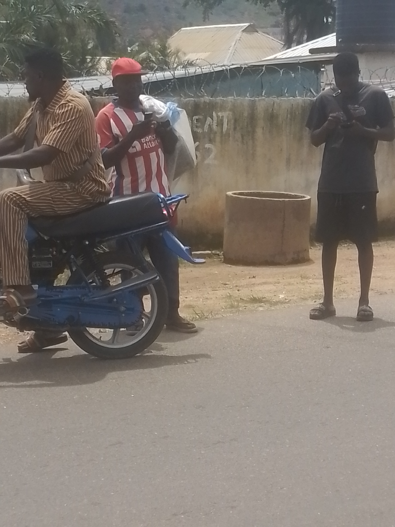 Three young men are on a street with a motorcycle. The man on the left is seated on the motorcycle, wearing a brown and white striped shirt and pants, and brown sandals. The man in the middle is standing next to the motorcycle, wearing a red and white striped shirt, a red baseball cap, and dark pants. He is holding a white plastic bag and a phone. The man on the right is standing a short distance away, wearing a dark t-shirt, black shorts, and dark sandals, and is looking at his phone. The scene appears to be outdoors in a somewhat rural or developing area, with a concrete wall with barbed wire in the background. There are also some buildings and vegetation visible. The lighting suggests it is daytime. The overall mood seems casual, with the individuals focused on their phones. Visible text includes "ENT" and "2" spray-painted on the wall.