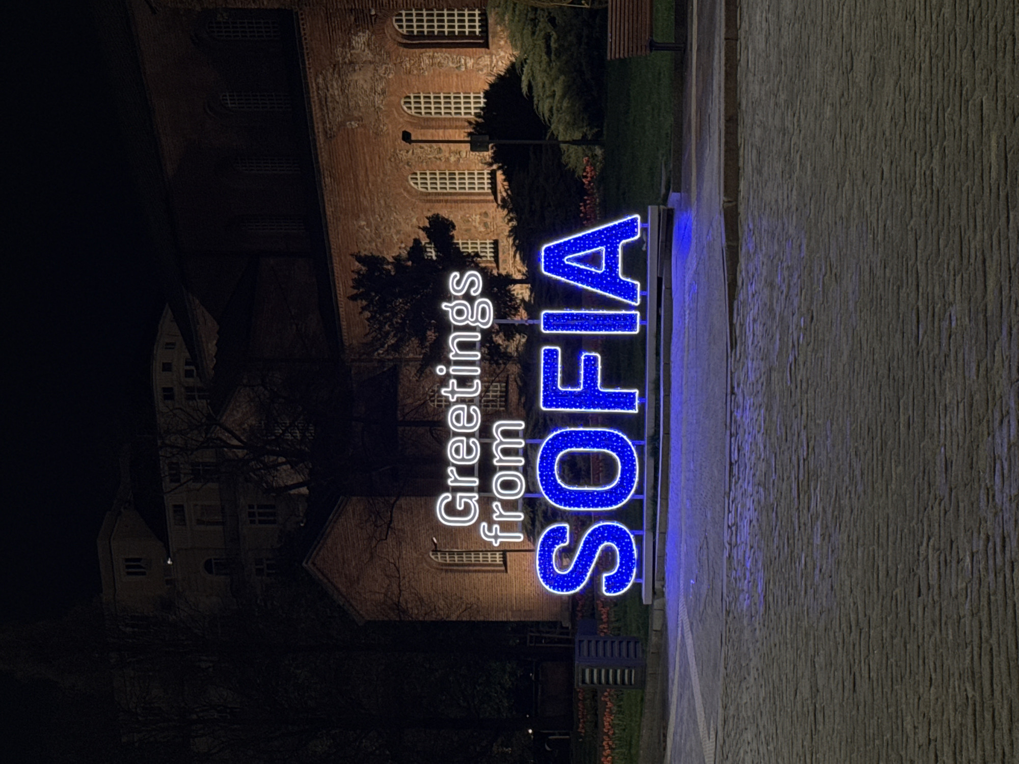 At night, a sign that reads "Greetings from SOFIA" in illuminated letters, with "Greetings from" in white and "SOFIA" in blue, is the focal point. The sign is located on a cobblestone sidewalk. Behind the sign is a brick building with arched windows. Trees and landscaping are also present. In the background, on the left, is another building, partially obscured by darkness and trees. The sign indicates that the location is in Sofia, Bulgaria.