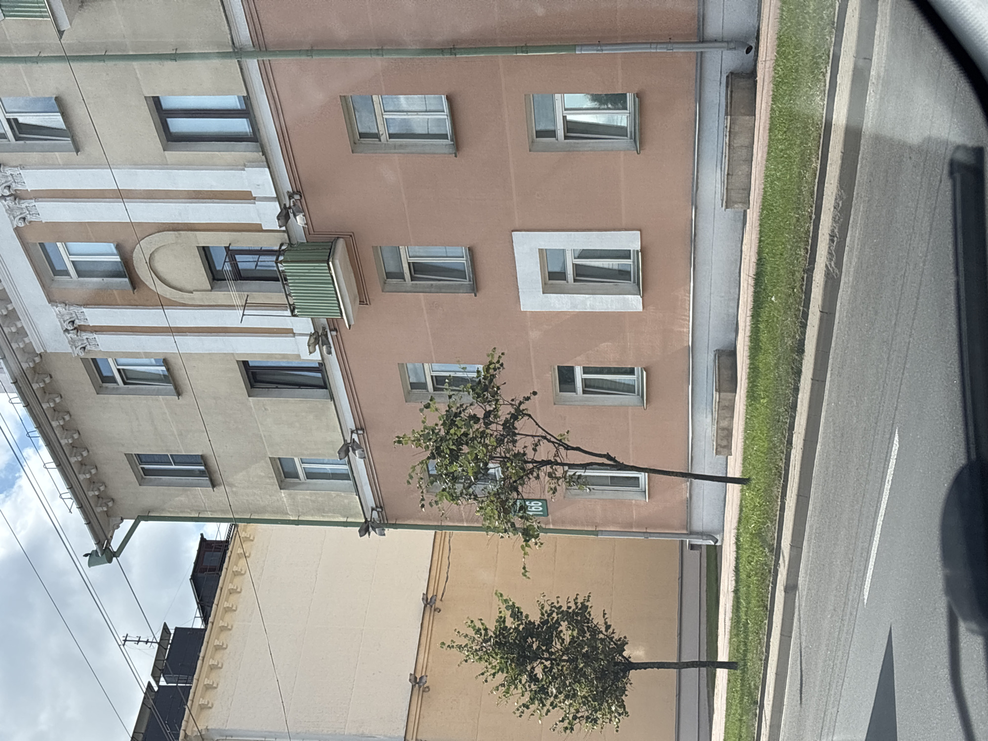 The image shows a building with a salmon pink facade and a pale yellow section on the left, located on a street in Minsk, Belarus. A street sign with the number "166" is visible on the salmon pink building. There are several windows on both sections of the building, some with white frames and some with dark frames. Two trees are planted on the sidewalk in front of the building. A road with a white line marking is in the foreground. The sky above is partly cloudy.