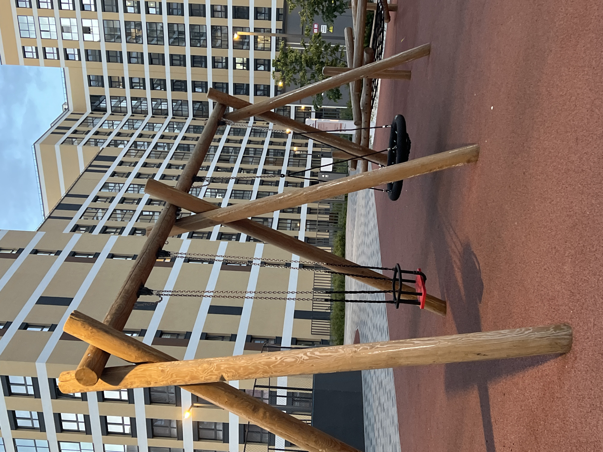 The image depicts a playground with a wooden swing set in the foreground, featuring one traditional swing with a red seat and chains, and a tire swing. In the background, multi-story residential buildings with beige facades and white trim are visible. The playground surface is a reddish-brown, cushioned material. The scene appears to be in a city environment, likely a residential area. There are no people visible in the image. The sky is overcast. Based on the architectural style of the buildings and the surrounding environment, this scene could be located in a modern urban development, such as a neighborhood in Yekaterinburg, Russia.