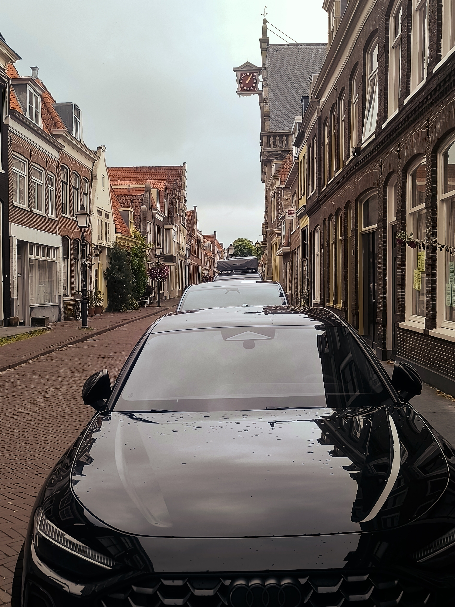This is a photograph of a street in the city of Hoorn, Netherlands. The street is paved with cobblestones and lined with historic brick buildings. In the foreground, a black car is parked, with its hood reflecting the surrounding architecture. Further down the street, other cars are visible, including a white SUV with a rooftop cargo carrier. A clock tower with a red clock face is visible on the left side of the street, part of an older building. The sky is overcast. The scene appears to be a quiet street, possibly in the morning or on a cloudy day.