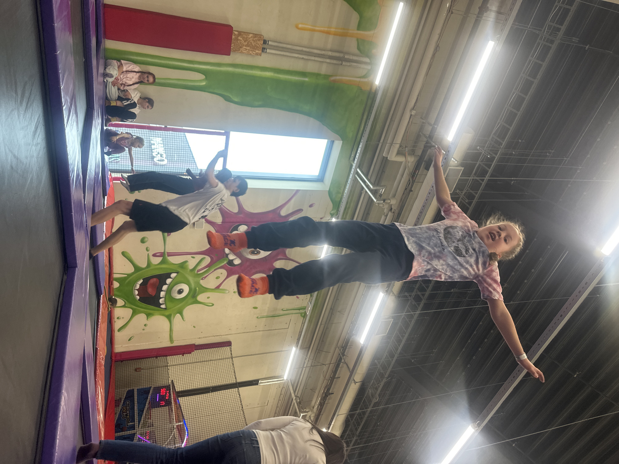 The image depicts the interior of a trampoline park. A young girl in an Ombre t-shirt and black pants with orange grip socks is in mid-air, seemingly jumping on a trampoline. Her arms are extended, and she is looking at the camera. Another child is visible further back, also in mid-air. A group of people are sitting along the side of a trampoline, watching the action. The trampoline is purple. The walls feature colorful graffiti-style art, including green and purple monster graphics. The ceiling is exposed, with metal supports and overhead lighting. The location of the trampoline park is Riga, Latvia.