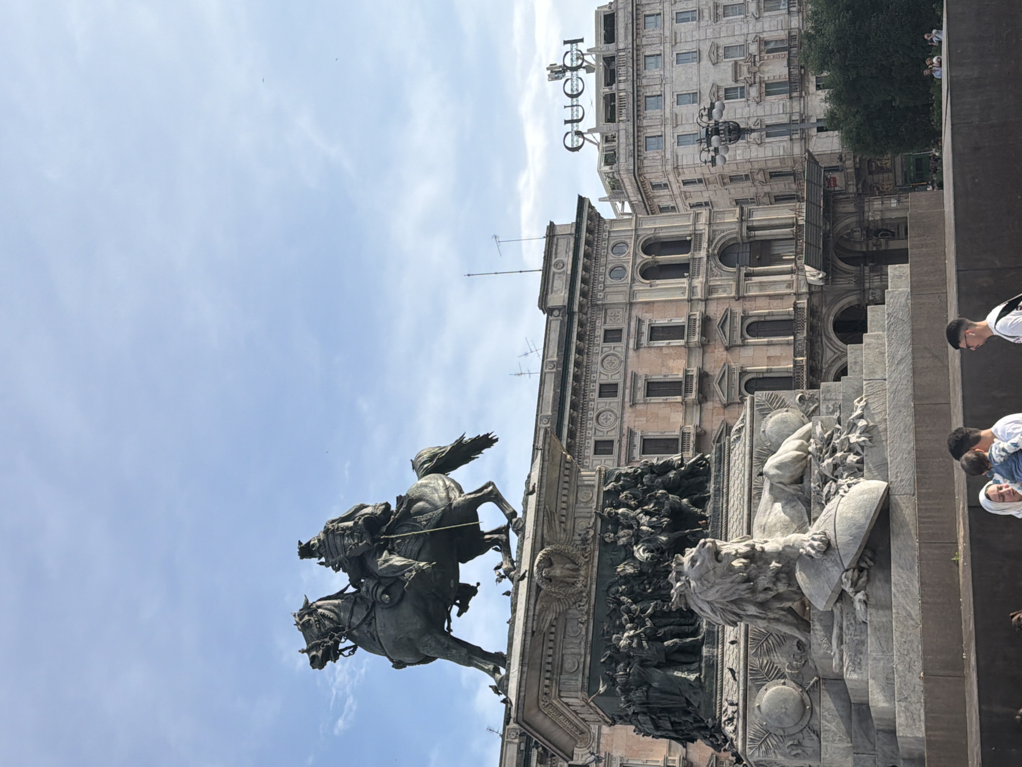 The image shows the equestrian statue of Vittorio Emanuele II in Piazza del Duomo, Milan, Italy. The statue depicts the king on horseback, atop a detailed pedestal adorned with sculptures of lions and figures. The Gucci sign is visible on the building behind the statue. Several people are nearby, with a group of tourists appearing on the right side. The sky is blue with some clouds.