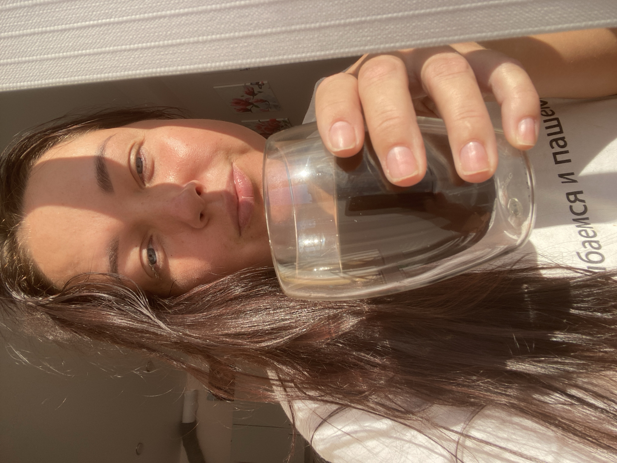A woman is holding a glass of dark liquid up to her face, partially obscuring her mouth. She has long, dark hair that falls across her shoulder and chest. She is wearing a white t-shirt with text printed on it, partially visible. The lighting suggests it is daytime, with sunlight casting shadows on her face and hair. The background shows a light-colored curtain or blinds on the right side of the frame and a light-colored wall on the left. There are also framed pictures of flowers on the wall in the background. The setting appears to be indoors, possibly a room in a residential building. The scene does not provide any specific visual cues to pinpoint a location within the city of Sochi, Russia.