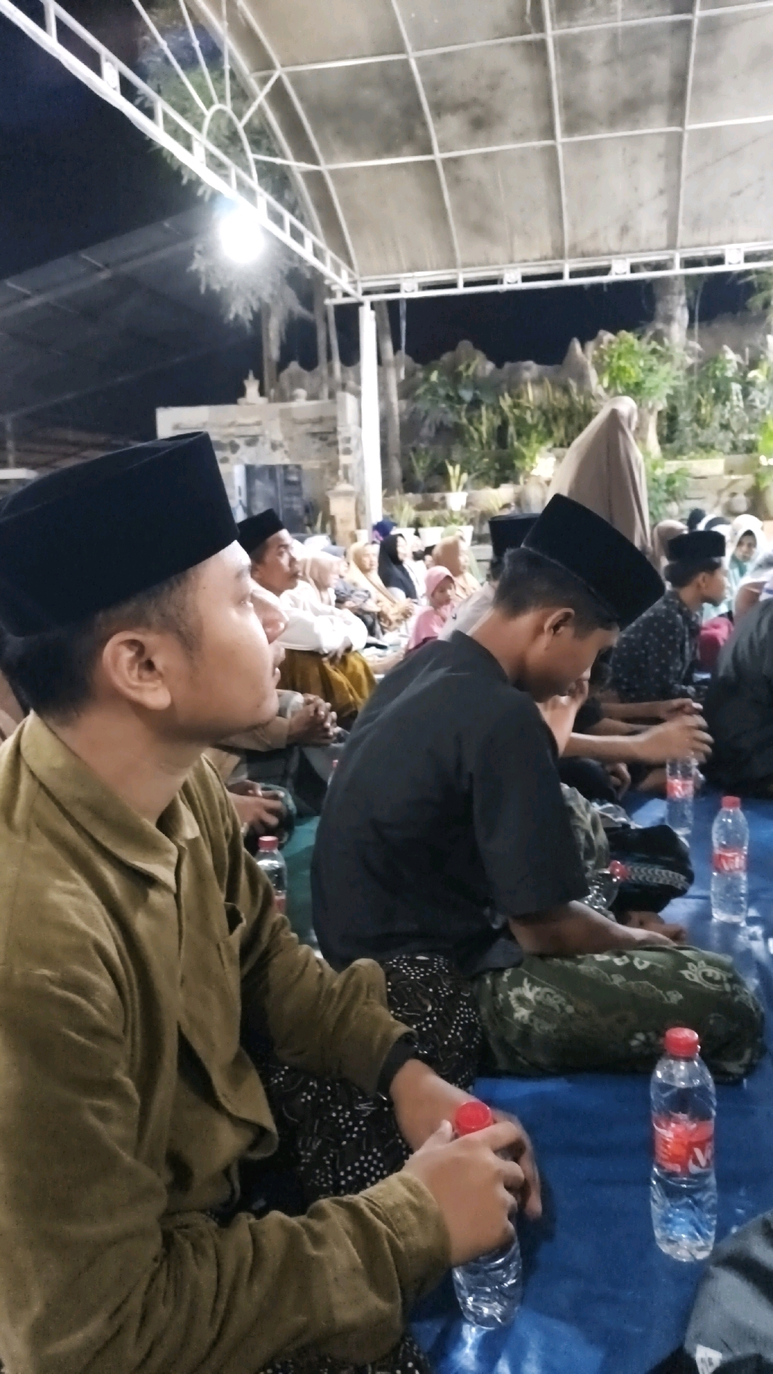 In Sambipondok, Indonesia, a group of people are gathered indoors, seated on a blue mat under a white canopy structure. The lighting suggests it is nighttime, with artificial lights illuminating the scene. In the foreground, a young man wearing a khaki shirt and a black peci (a type of cap) is seated on the left, facing the right. Next to him, a young man in a black shirt and patterned pants is also seated, facing towards the front. Several other individuals, including men, women, and a child, are visible in the background, also seated on the mat. Some individuals are wearing peci caps and headscarves, indicating a religious or cultural gathering. Several bottles of water are placed on the mat in front of the seated individuals. The environment appears to be an outdoor or semi-outdoor gathering space, with some greenery and structures visible in the background.