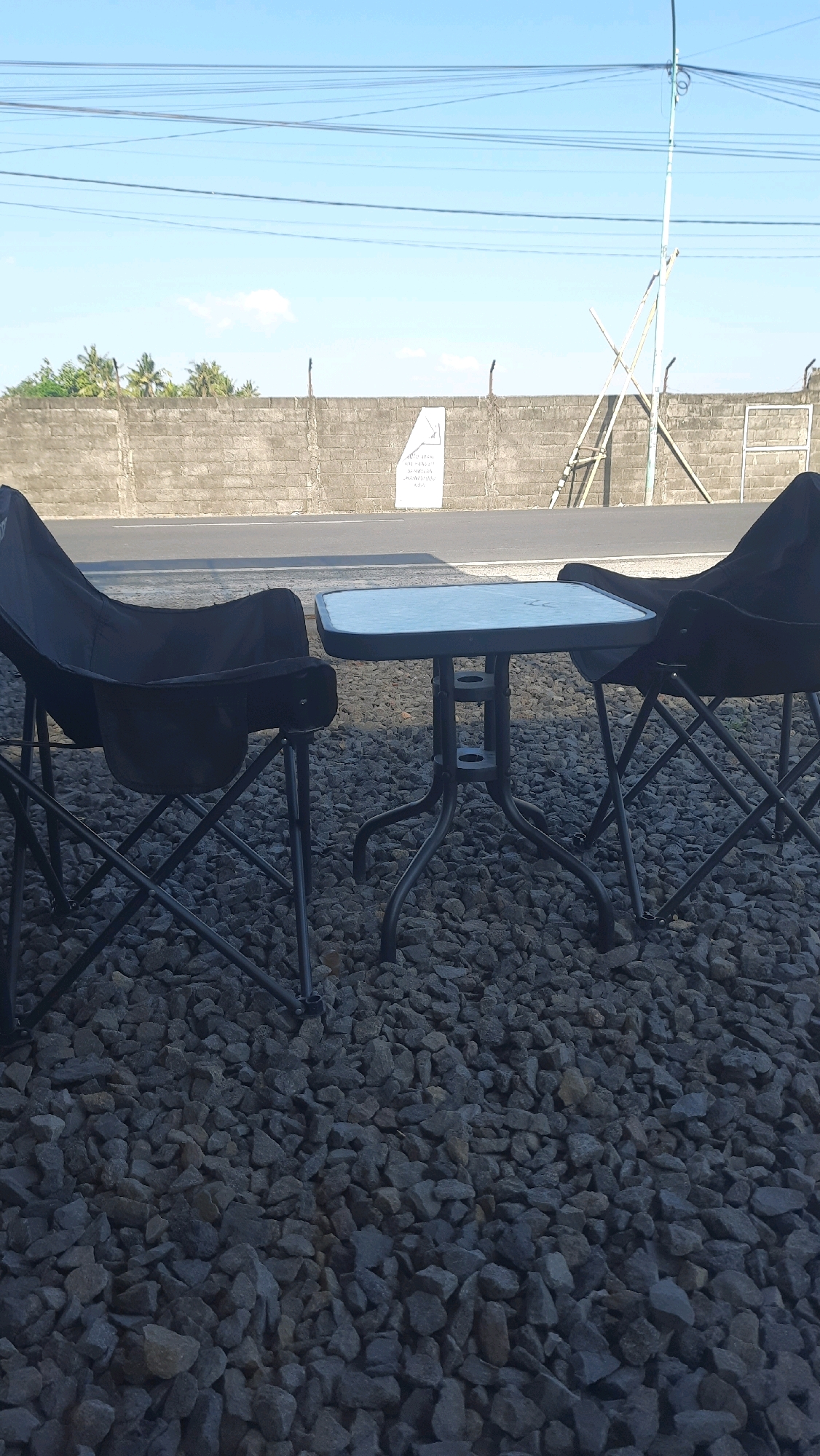 The image shows an outdoor seating area with two black folding chairs and a small square table set on a bed of gray gravel. The chairs and table are positioned in front of a grey brick wall. Beyond the wall, there is a road and a strip of vegetation, likely palm trees, under a clear blue sky with some power lines overhead. The scene appears to be in Jajag, Indonesia, possibly a rest stop or a roadside cafe.
