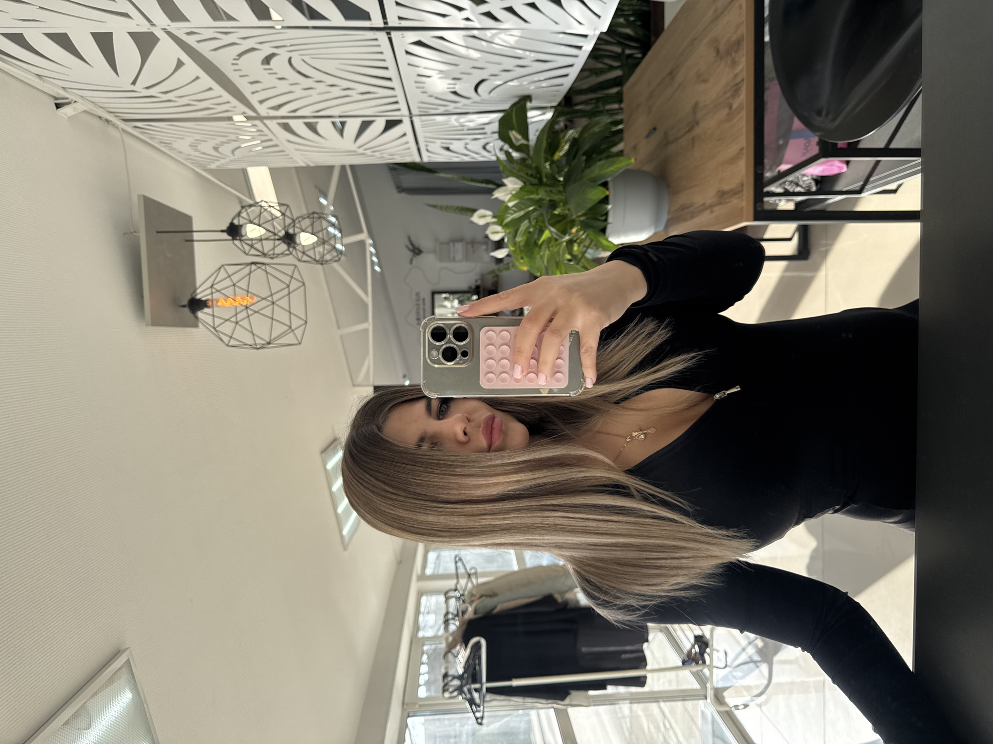 A young woman is captured in a mirror selfie taken indoors in Chaikovsky, Russia. She has long, straight hair with light brown and blonde highlights, and wears a black long-sleeved top paired with a delicate gold necklace featuring a small pendant. Her expression is neutral, with slightly pursed lips. She holds an iPhone-like smartphone with a transparent case, which has a pink pop-it toy integrated into its back, partially obscuring her face.
The reflection in the mirror reveals a bright, modern interior, indicating it is likely daytime due to the natural light streaming in. The setting features a white ceiling or decorative wall panel adorned with intricate, geometric cut-out patterns. Three contemporary black wireframe light fixtures, each containing a warm, amber-glowing filament bulb, are visible below the patterned panel. A lush green plant, identified as a Peace Lily with white flowers, rests in a simple grey pot on a wooden surface, possibly a table or countertop. Further in the background, a black metal clothes rack holds various garments, positioned in front of a window that emits bright light. The walls exhibit a subtle horizontal textured finish. The overall ambiance suggests a stylish and well-lit space, possibly a salon, studio, or commercial establishment. No legible text is visible in the image.