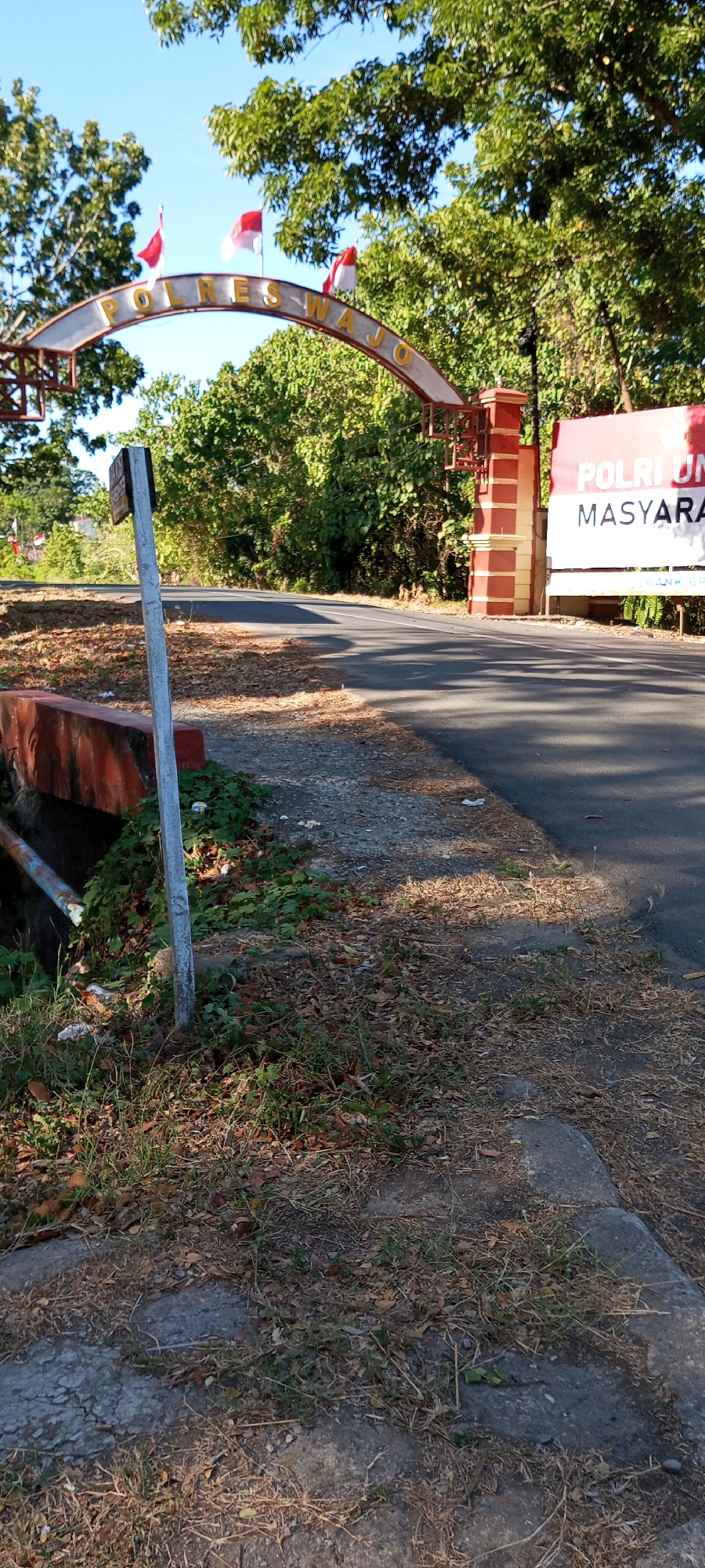 An outdoor, eye-level, full shot captures a paved road winding through a vegetated area under a clear blue sky. In the foreground, a gray pole stands to the left of the road. To the left of the pole is a concrete barrier with orange accents, and beyond it, green foliage. To the right of the road, a portion of a large white banner with red lettering, displaying "POLRI UMUM MASYARAKAT," is visible, situated behind a red and yellow striped pillar. An arched entranceway with the words "POLRES WAJO" in gold lettering spans over the road, adorned with three Indonesian flags. The ground beside the road is covered with dry leaves and sparse green grass. The scene appears to be the entrance to the Polres Wajo in Bulupabbulu, Indonesia.
