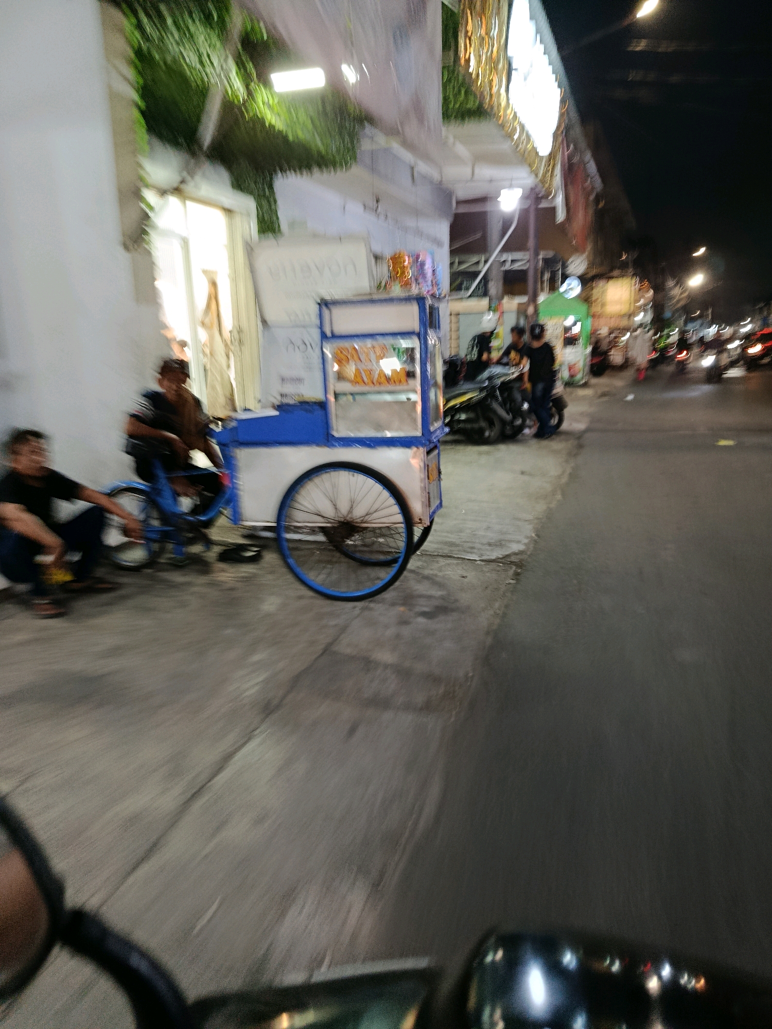 The image depicts a street scene at night in Bekasi, Indonesia, characterized by motion blur from movement, likely from a vehicle. In the foreground, a blue and white food cart is parked on a sidewalk. Two individuals are seated near the cart; one is on a blue bicycle and the other is crouching on the ground. Further down the street, several motorcycles are visible, some with riders. The street is illuminated by overhead lights and the headlights of passing vehicles. Buildings line the street, some featuring illuminated signs. Green foliage cascades from an overhang above one of the buildings. The overall impression is of a busy urban environment at night.