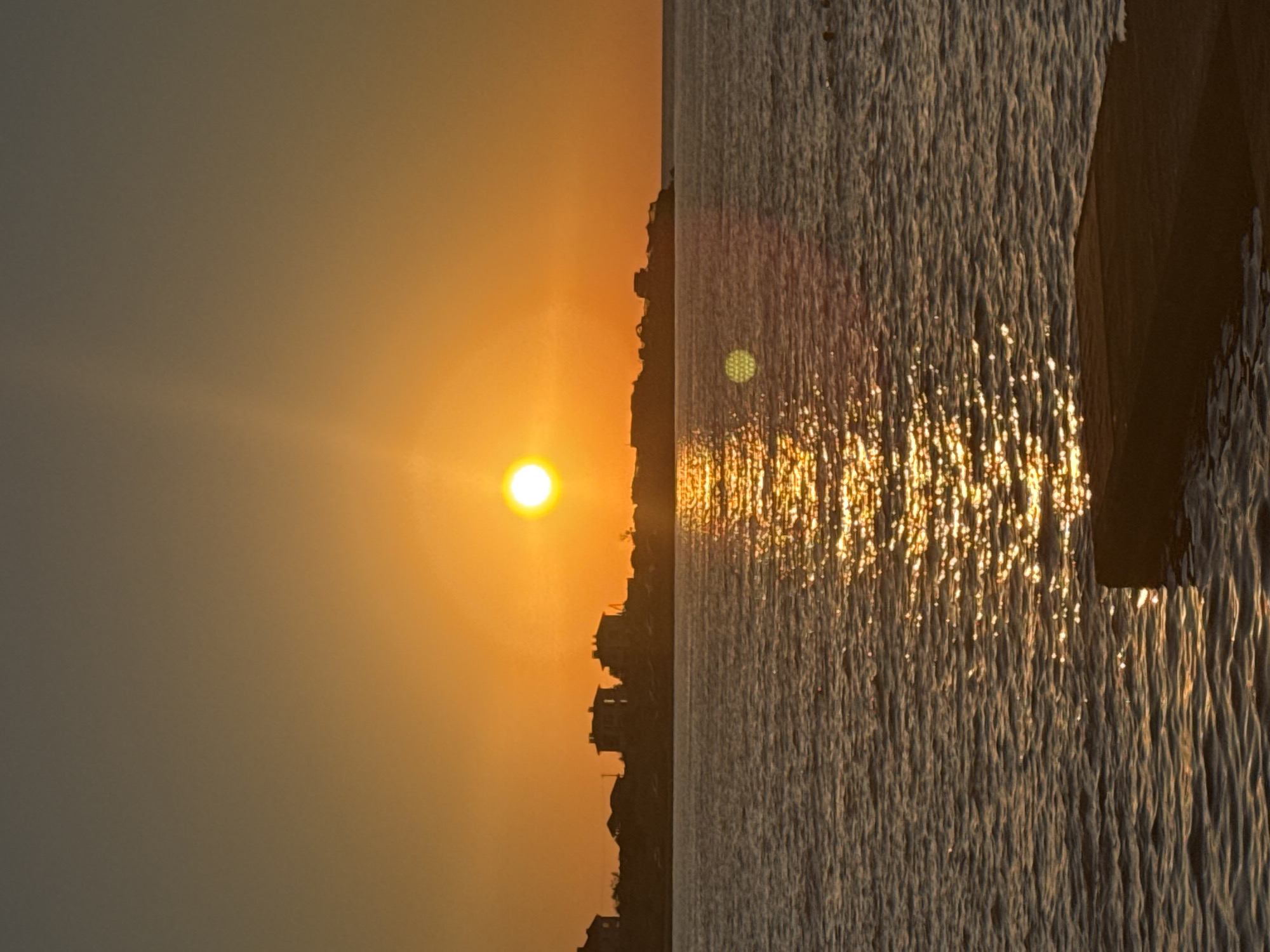 A vertical shot captures a sunset over the Black Sea in Sevastopol, Ukraine. The sun, a bright orb, hangs in the orange sky, casting a shimmering golden path across the water. Silhouetted against the warm glow are buildings and structures lining a distant shore. In the foreground, a dark, rectangular object, possibly a pier or a submerged structure, breaks the surface of the water. The sea is calm, with gentle ripples reflecting the light. The sky transitions from a deep orange near the horizon to a dusky gray at the top of the frame.