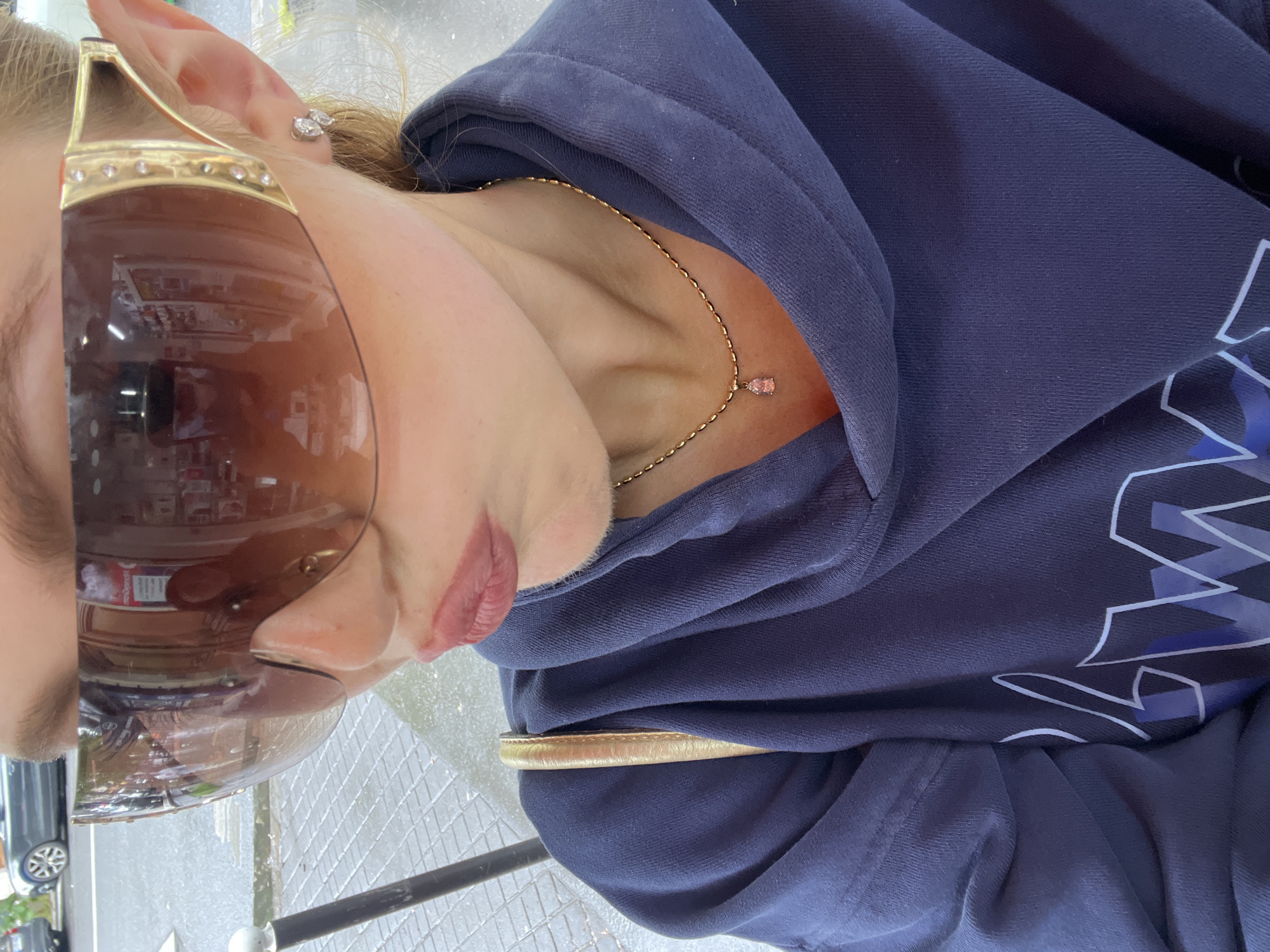 A vertically-oriented, medium close-up photograph shows a woman wearing dark sunglasses, a gold necklace with a small pink stone, and a dark blue hoodie with a white and blue design. She is wearing small earrings. The reflection in her sunglasses shows what appears to be the interior of a store with shelves of products. Outside the store is a street with cars parked along it. The location is implied to be Paris, France, due to the presence of signage in French and the general architectural style visible in the reflections. The ground appears to be pavement or a sidewalk. There is a metal post with a white top in the foreground.