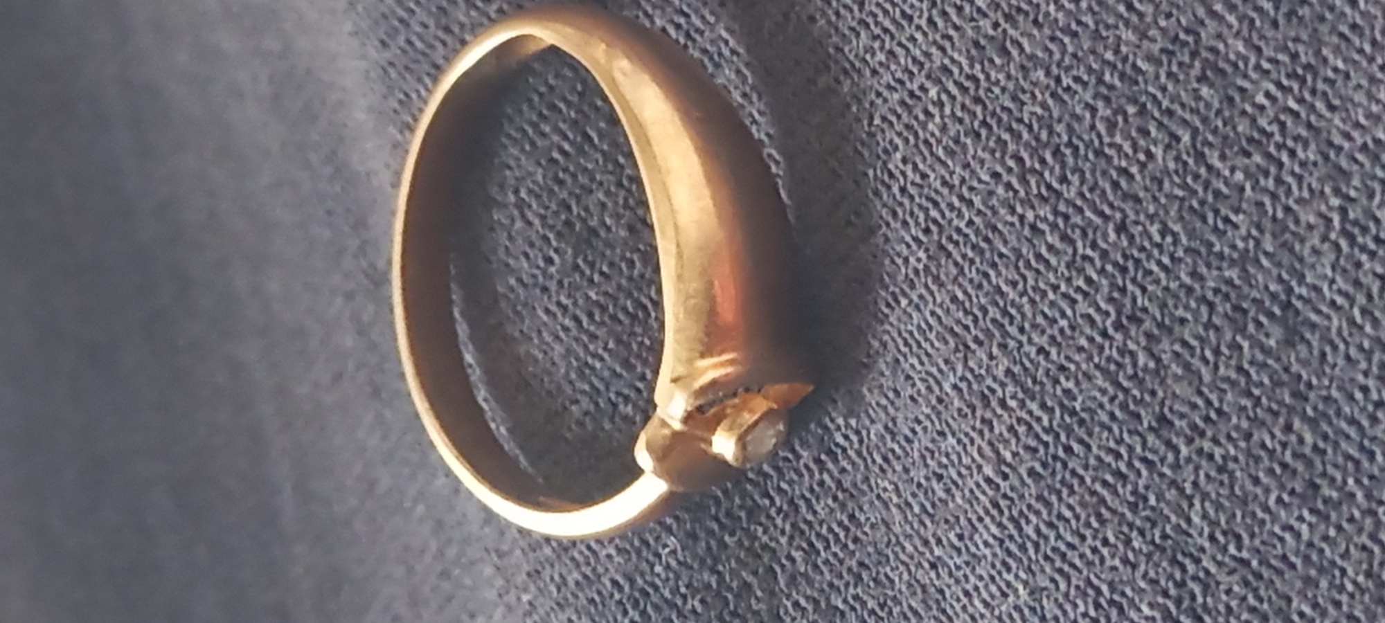 A gold ring with a single stone is lying on a textured, dark blue or gray fabric. The ring's band is smooth and curved, leading to a setting that holds a small, possibly faceted, stone. The background fabric shows a uniform, slightly rough texture.
The location or any reference to the city of Kryvyi Rih, Ukraine cannot be deduced from the image.