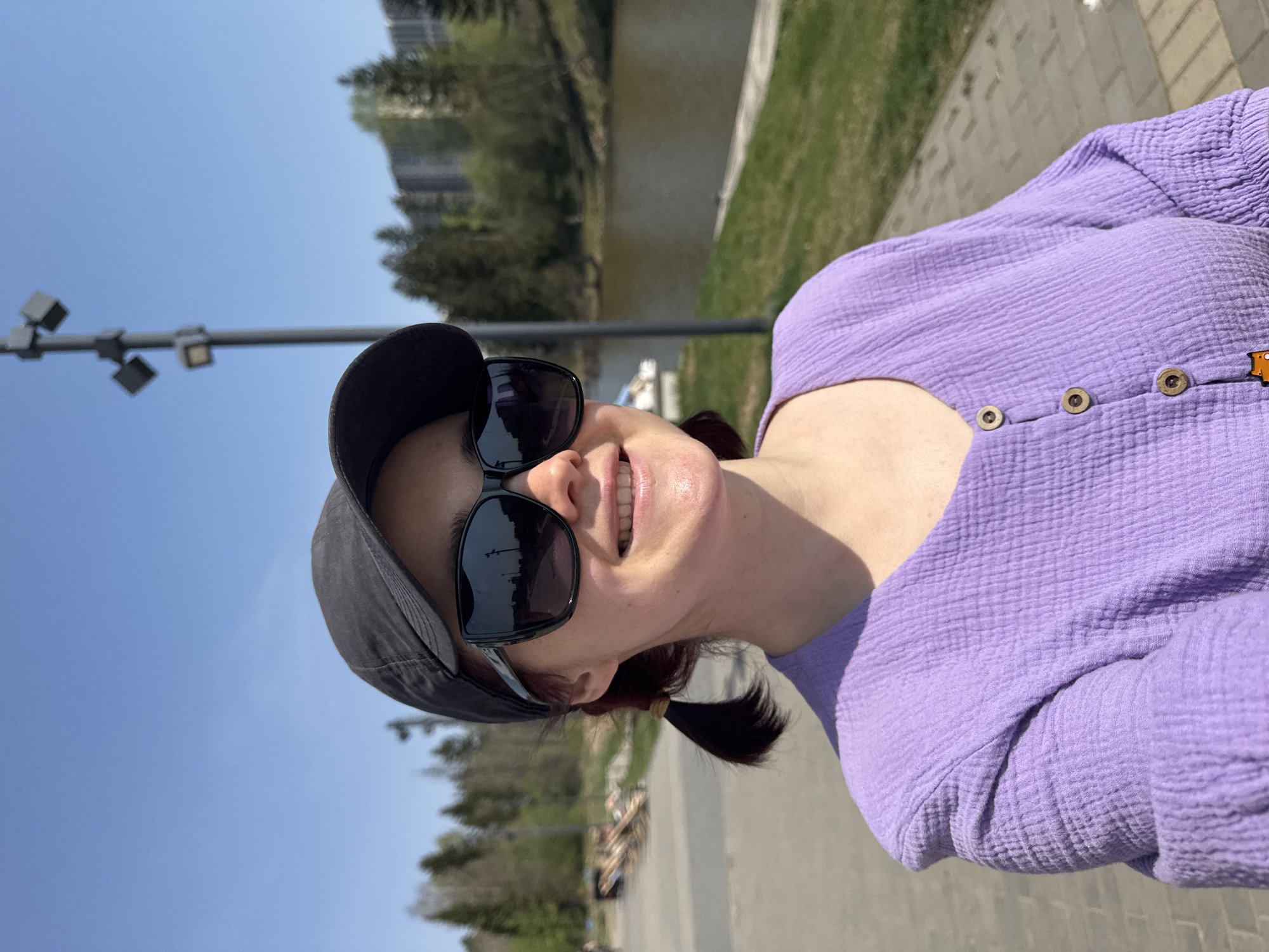 Here is a description of the image:
The image is a selfie of a woman with light skin wearing a purple textured shirt with wooden buttons, black sunglasses, and a dark-colored baseball cap. She has dark hair pulled back into a ponytail. The background reveals an urban park setting with a river or canal, trees, a street lamp, and a paved walkway. The location is likely a public space in the city of Izhevsk, Russia.