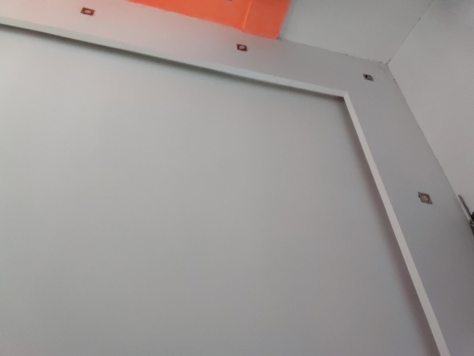 This media file presents an interior view of a room, likely located in Santa Ana, Venezuela. The central subject is a large, light grey wall featuring a prominent white architectural trim. This trim forms a partial rectangular frame, running along the top and right edges of the visible wall section.
Embedded at regular intervals within the white trim are several small, dark, square or rectangular cutouts or fixtures. These elements are visible along the horizontal upper trim and the vertical trim on the right side. Above the top white trim, an orange-colored section is prominent, suggesting either a ceiling painted orange or an upper wall segment with a distinct color.
The image shows a corner of the room on the right, where the light grey wall meets another wall, also appearing in a light tone. A small, dark object, possibly a bracket or part of a mounted device, is partially visible near the top right corner of the image.
No people are present, and no specific activities are taking place. The scene appears static, showcasing architectural details of what might be a contemporary or newly constructed/renovated space. The lighting is uniform, suggesting an indoor setting without indicating a particular time of day or specific weather conditions. There is no visible text within the image.