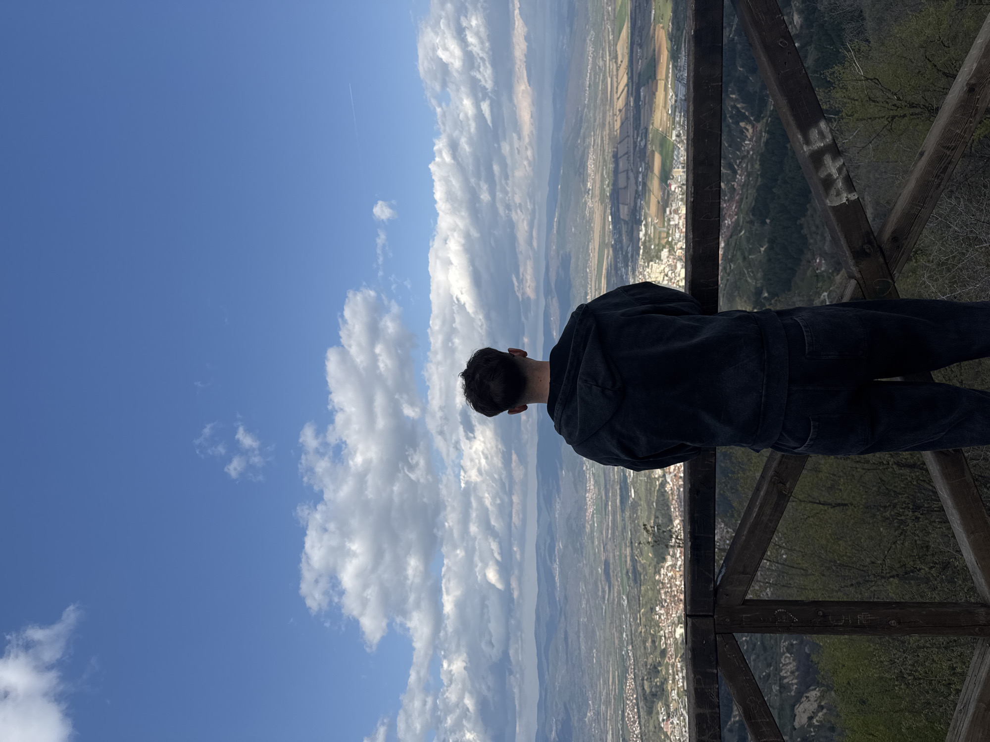 The image shows a person standing on a wooden observation deck, gazing out at a landscape. The person is wearing a dark-colored jacket and dark pants, and only the back of their head and upper body are visible. The observation deck is made of wooden planks, forming a railing. Beyond the railing, there's a panoramic view of a valley with scattered buildings and green patches of land, suggesting a rural or suburban area. The sky takes up a large portion of the frame, featuring a gradient of blue with puffy white clouds. The location appears to be Delchevo, Bulgaria, as suggested by the landscape and built environment visible in the background.