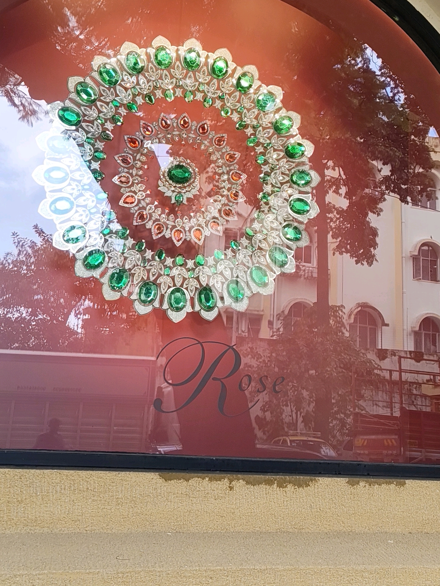 The image shows a reflection in a shop window. In the foreground, a large, ornate necklace with green and orange gemstones is displayed against a red background. Behind the necklace, a reflection of a building and trees can be seen. A taxi and a truck are also visible in the reflection. The building appears to be a multi-story structure with arched windows. The overall scene suggests an outdoor setting in a city, likely during the daytime. The text "Rose" is visible in the reflection of the shop window, indicating the name of the establishment. The specific location in Mumbai, India, cannot be determined from the image alone, but the presence of a taxi and a truck suggests an urban environment.