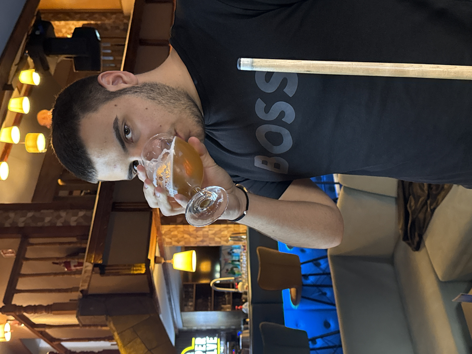 A man wearing a black t-shirt with "BOSS" printed on it is drinking from a glass containing a beverage. He has short dark hair and a beard. A wooden cue stick is positioned horizontally across his body. The background reveals an interior setting with a bar area featuring bar stools and blue ambient lighting. The location seems to be in Plovdiv, Bulgaria, based on architectural design and lighting fixture styles.