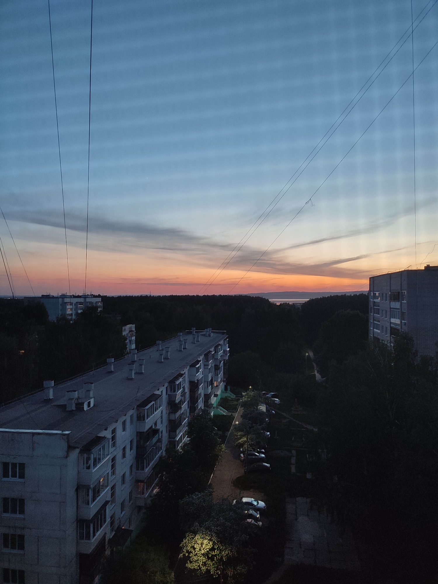 The image depicts a cityscape at dusk. In the foreground are multiple apartment buildings, characterized by gray concrete exteriors and rectangular structures. Behind the buildings, there is a dense forest. In the background, the sky transitions from a light blue to a deep orange and red, suggesting either sunrise or sunset. Utility lines are visible across the sky. The lighting is dim, with some artificial lights illuminating sections of the ground and trees. The image likely represents a residential area on the outskirts of a city, given the combination of apartment buildings and forest. The location is noted as being in the city of Unknown, Unknown. There is no visible presence of people, but there are parked vehicles in the middle ground.
