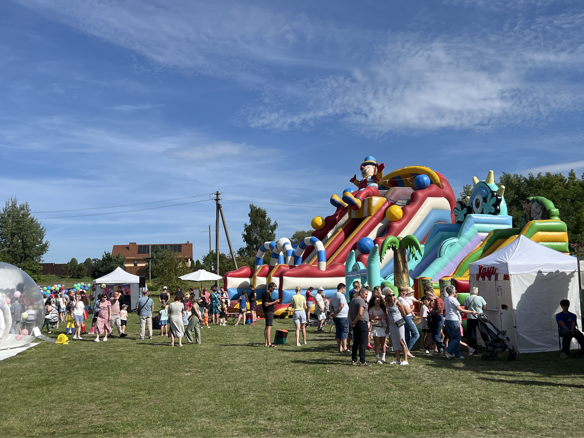 In Vilnius, Lithuania, on a sunny day, a vibrant outdoor event unfolds on a grassy field. The centerpiece is a large, inflatable obstacle course with multiple slides and climbing sections, adorned with colorful cartoon characters like a clown and a monster. A crowd of people, varying in age and attire, mingles in front of and around the inflatable attraction. Some are dressed casually in t-shirts and shorts, while others opt for dresses and more formal wear. Children are present, some appearing to be in groups, suggesting families enjoying the festivities.
Several temporary structures are set up, including white tents that may serve as information booths or vendor stalls. One tent has text that appears to read "Kasa," likely indicating a ticket or payment point. A white umbrella offers shade near one of the tents. In the background, trees form a natural backdrop, and residential buildings with brown roofs are visible, suggesting the event is taking place in a suburban or park-like area. Power lines and utility poles are also present, indicating the proximity of infrastructure.
The ground is covered in green grass, and a clear blue sky with scattered white clouds presides over the scene. The atmosphere appears festive and relaxed, with people engaged in conversation, standing in lines, or simply observing the activities. The overall impression is one of a community gathering or a local festival, likely aimed at families with children, capitalizing on good weather for an outdoor celebration.