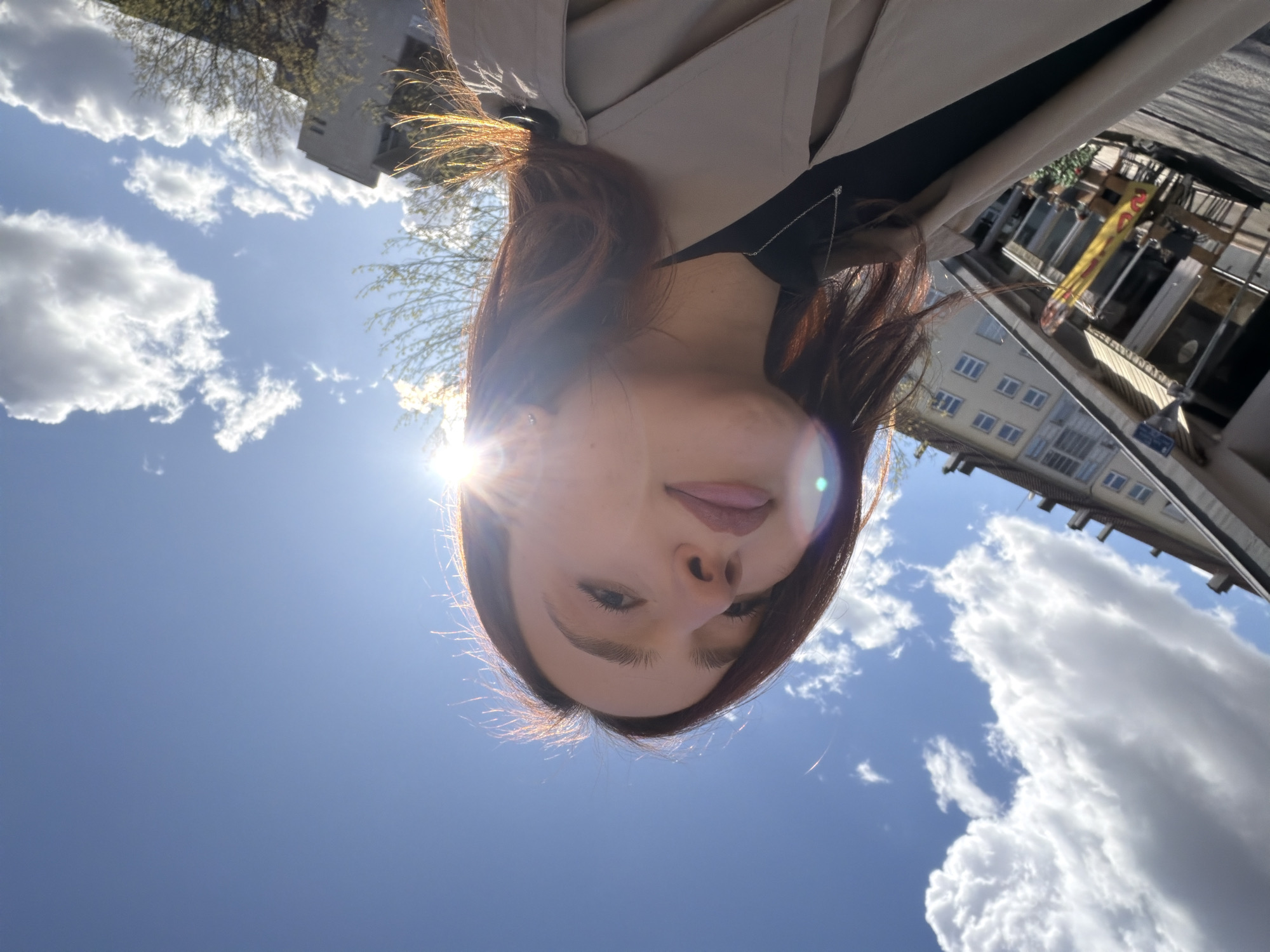 A young adult female is depicted in a low-angle, sunlit selfie. Her face, positioned centrally, is angled upwards and slightly to the viewer's right, revealing light-colored lips, dark eyebrows, and brown hair backlit by intense sunlight, which creates a lens flare across her upper face and right cheek. She wears a black top beneath a beige trench coat, accented by a delicate silver chain necklace with a small pendant.
The scene is an urban streetscape in Strasbourg, France, specifically on **Rue de la 1ère Armée**. To the left of the subject, a multi-story building with a light-colored facade, numerous windows, and a distinctive top floor featuring enclosed balconies is visible. Adjacent to this building, a commercial establishment, likely a cafe or restaurant, features outdoor seating with tables and chairs beneath a striped awning. A yellow banner with red, stylized text is positioned near this establishment's entrance. Further into the background on the right, additional buildings and trees displaying early spring foliage are present. The sky is clear and bright blue, interspersed with scattered white cumulus clouds, indicative of sunny weather.
