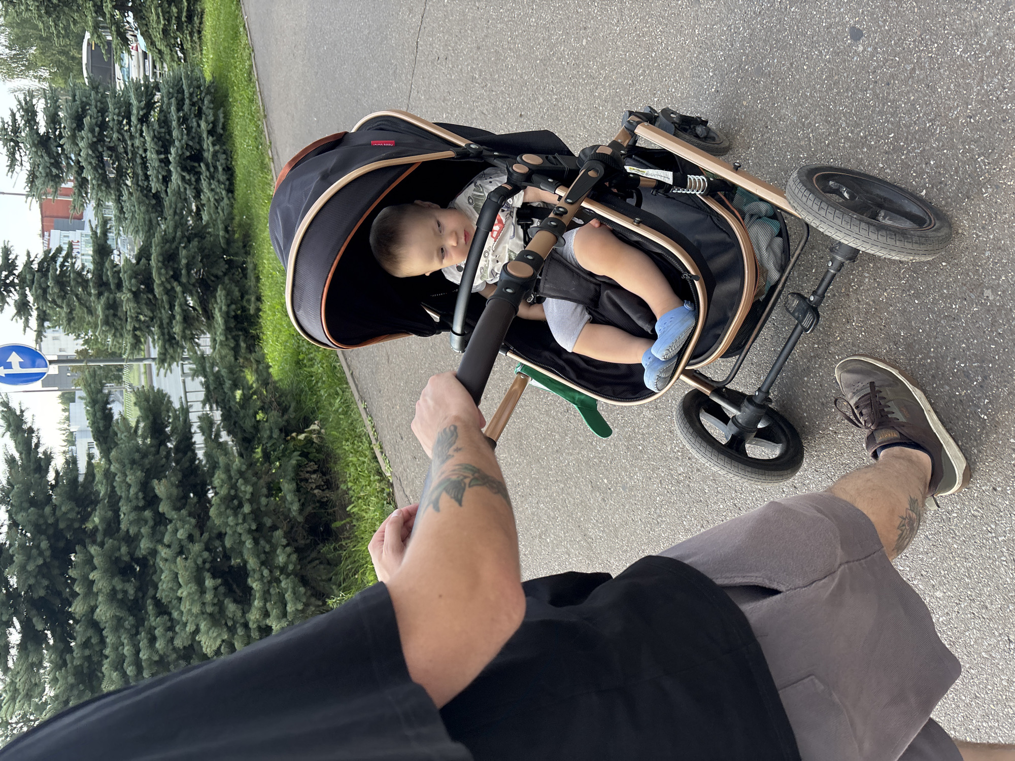 A close-up, eye-level shot shows a person's legs and hands pushing a stroller with a baby. The person is wearing a black t-shirt and gray shorts, and has tattoos on their arm and leg. The stroller is black and gold, with a baby sitting inside. The baby is wearing a patterned t-shirt and blue shoes. The scene takes place outdoors on a paved path, with green trees and a sidewalk in the background. There is a traffic sign with a blue circle and white arrow in the upper left corner of the image, suggesting this is a public area in Naberezhnye Chelny, Russia. The overall lighting indicates it is daytime.