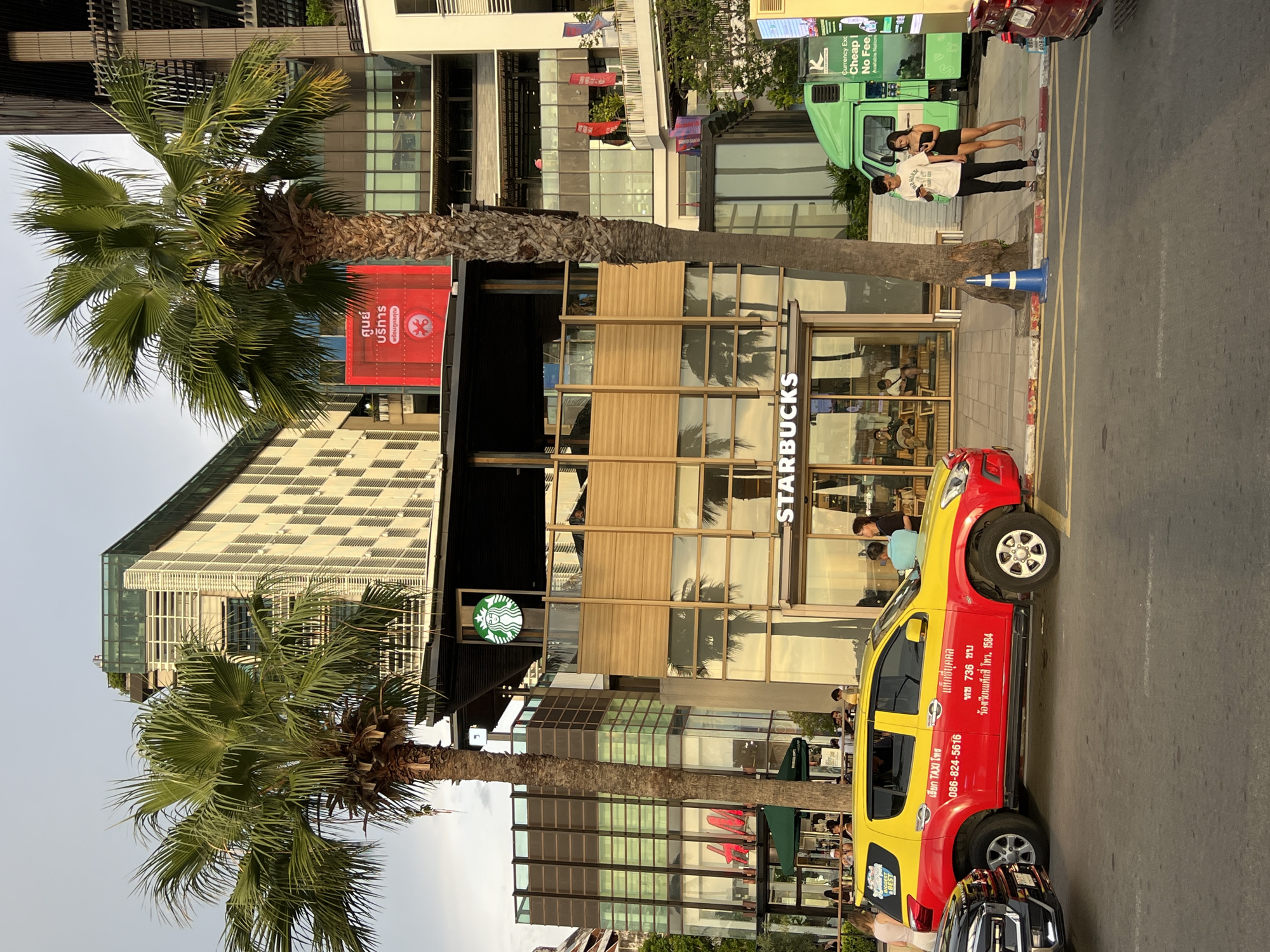 A vibrant street scene in Pattaya City, Thailand, unfolds under a bright sky. In the foreground, a distinctive red and yellow taxi, an SUV model, is parked alongside a black car. The taxi is adorned with Thai script and phone numbers, and a sticker on its rear window declares "BEST". Further down the street, a bright green delivery truck with the text "Currency Exchange Cheap No Fee Available Nationwide" on its side is visible. Two young adults, a man and a woman, stand near the truck, both engrossed in their mobile phones.
The backdrop is dominated by a modern, multi-story building featuring a Starbucks prominently displayed on the ground floor. The Starbucks facade is characterized by large glass windows and wooden accents. Two large palm trees stand tall, their fronds reaching towards the sky, adding a tropical flair to the urban landscape. Reflections of palm trees and the sky are visible in the glass windows of the building. Beyond the Starbucks, the architecture becomes more complex, with various levels and facades, including a checkered pattern on one section of the building. Several people are seen within the Starbucks and on the outdoor seating area, enjoying their time. The street itself is paved, with yellow and white lines marking parking spaces and traffic lanes. A blue traffic cone stands near a palm tree on the sidewalk.