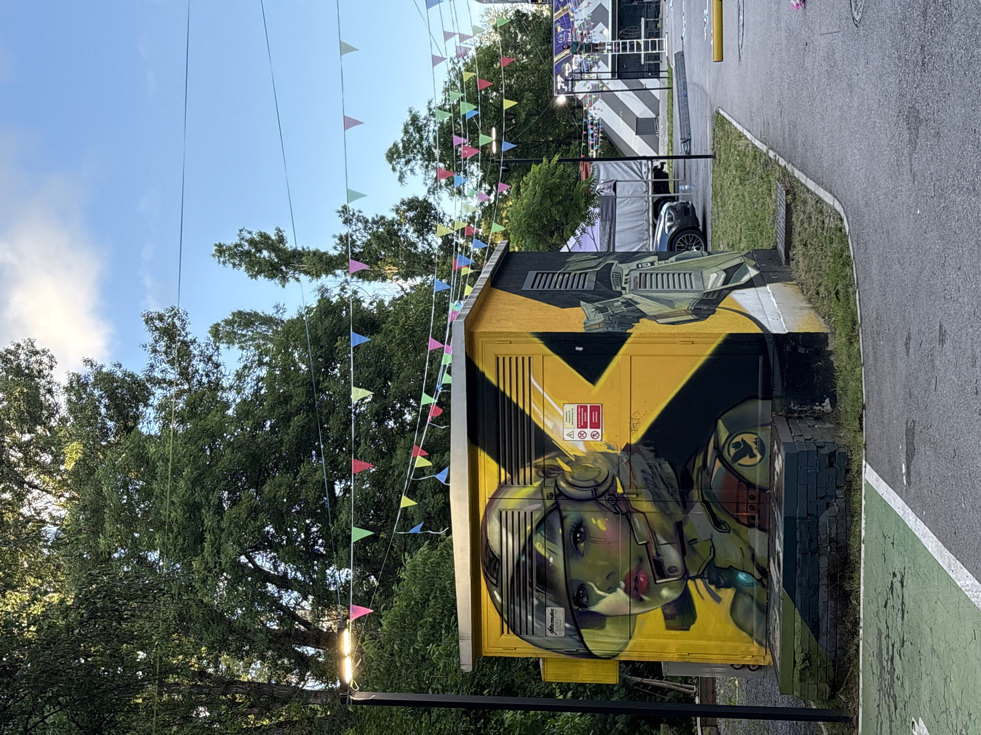 This image depicts a vibrant outdoor scene, likely in Sofia, Bulgaria, featuring a colorfully graffitied utility box. The utility box is painted bright yellow with black accents, and its main feature is a large, detailed graffiti artwork of a female figure. This figure, possibly an astronaut or pilot, is shown from the chest up, wearing a futuristic helmet or headset, with prominent red lips and expressive eyes. The art incorporates shades of green and gray for the figure and helmet, set against the yellow and black of the box. Above the figure, industrial-looking machinery or structures are also painted onto the box.
Several warning signs are visible on the yellow surface of the utility box, including a lightning bolt symbol, a "danger" symbol, and the word "ОПАСНО!" (Bulgarian for "Dangerous!"). A small signature, possibly "Memurks," can be seen near the figure's helmet. A symbol resembling a bird or phoenix is also painted on the figure's shoulder.
Above the utility box and extending across the upper portion of the frame, numerous strings of colorful triangular flags (bunting) are suspended, suggesting a festive or celebratory atmosphere. Lush green trees with dense foliage frame the left and upper parts of the image, indicating a park or heavily vegetated area.
To the right, an asphalt path or road is visible, featuring a green-painted section and a white line, possibly designating a bike lane or pedestrian path. A strip of grass and a concrete curb separate the path from the utility box. In the background, a dark car is partially visible, along with a building or event structure characterized by a distinctive black and white striped pattern. The scene is bathed in bright daylight under a clear blue sky with some scattered white clouds.