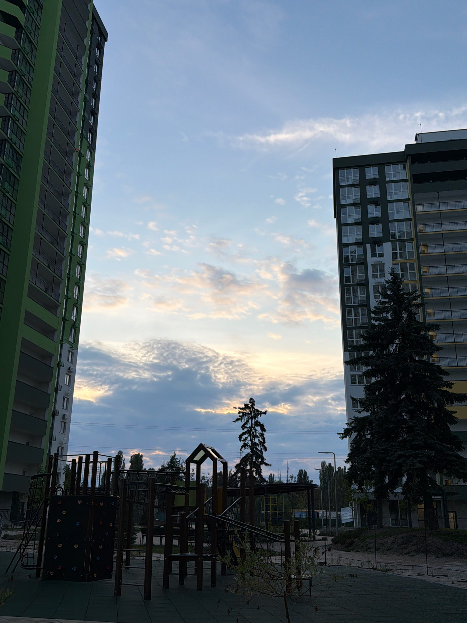 The image depicts a children's playground in front of two apartment buildings, with a view of a cloudy sky at dusk. The building on the left is tall and predominantly green, with balconies and numerous windows. The building on the right is shorter and has a darker facade, also featuring balconies and windows, with a large pine tree in front of it. The playground equipment, made of dark wood and metal, includes a climbing wall, swings, and slides. The ground of the playground is a dark rubber mat. Several trees are visible in the background, as well as streetlights and power lines. The sky displays a mix of light and dark clouds, with hints of orange and yellow suggesting the setting sun. The scene is set in a residential area of Kyiv, Ukraine. There are no people visible in the image.