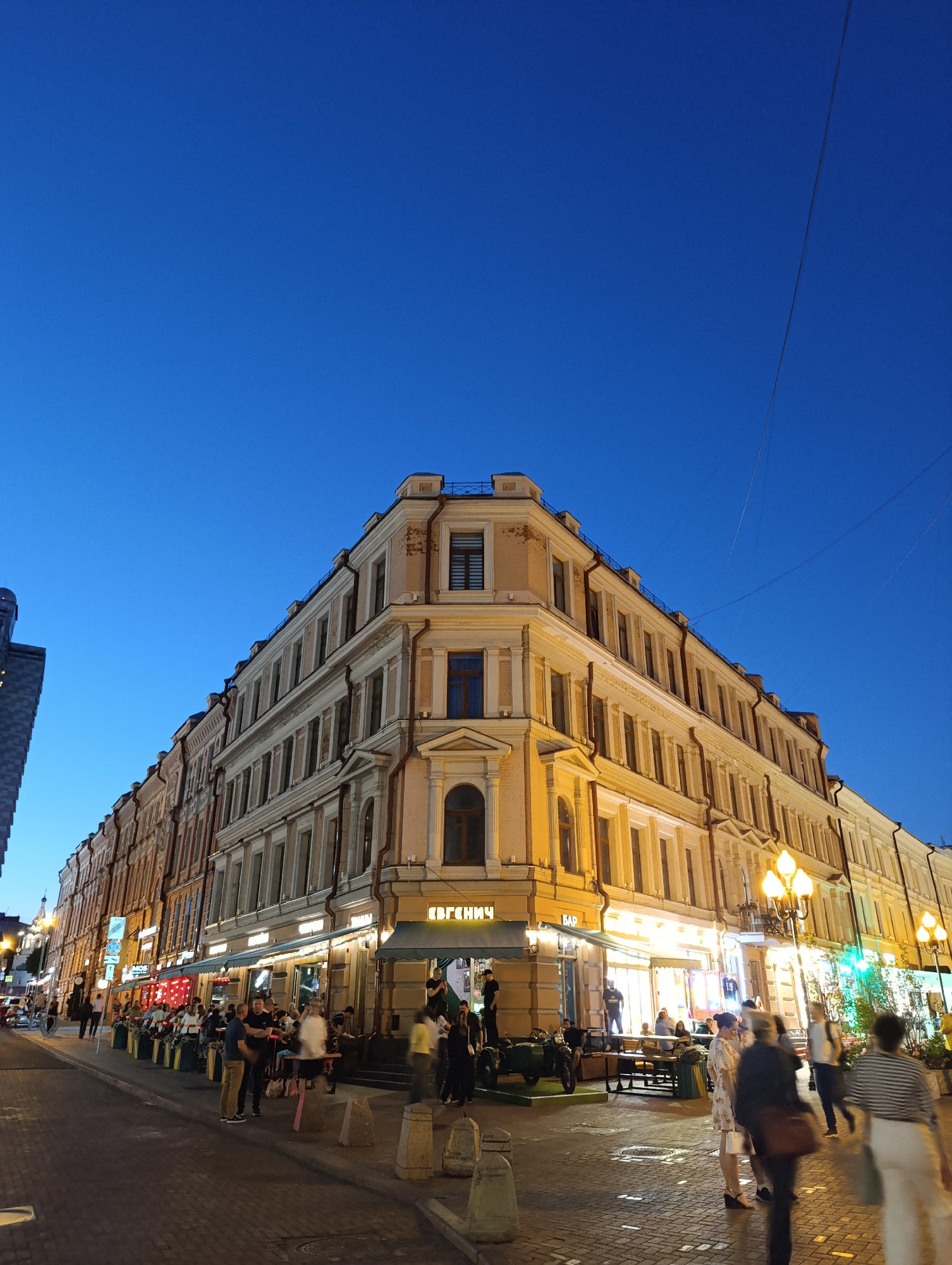 This image depicts Rozhdestvenka Street, 19/2, in Moscow, Russia, during twilight. The sky is a deep, clear blue, indicating early evening after sunset. Streetlights and building illuminations are active.
The central focus is a five-story, cream-colored historic building with classical architectural details, including cornices, decorative window frames, and balustrades on the roofline. It occupies a prominent street corner. The ground floor of this building houses commercial establishments, notably the "Евгенич" (Evgenich) Bar on the corner, identified by Cyrillic signage and green awnings.
To the left of the corner building, a row of multi-story buildings extends down a street, some with similar light facades, others appearing to be reddish-brown brick. Signs, including "MOS," are visible on these buildings. A modern, dark-colored building is partially visible on the far left, contrasting with the historical architecture. A distant church dome or steeple is faintly visible further down the street on the left.
To the right of the corner building, the street continues with similar multi-story historic buildings, also featuring illuminated ground-floor businesses and outdoor seating areas. Ornate, multi-lamp streetlights illuminate this side of the street. Overhead wires are visible against the sky.
The street level is paved with dark cobblestones or pavers. Numerous people populate the scene, indicating a lively atmosphere. Many individuals are seated at outdoor tables and chairs, arranged in front of the establishments, dining and socializing. Other people are seen walking on the pavement, some exhibiting motion blur due to movement. Concrete bollards are placed along the edge of the pedestrian zone. A blue circular road sign with a red diagonal bar is visible on the left side of the street.