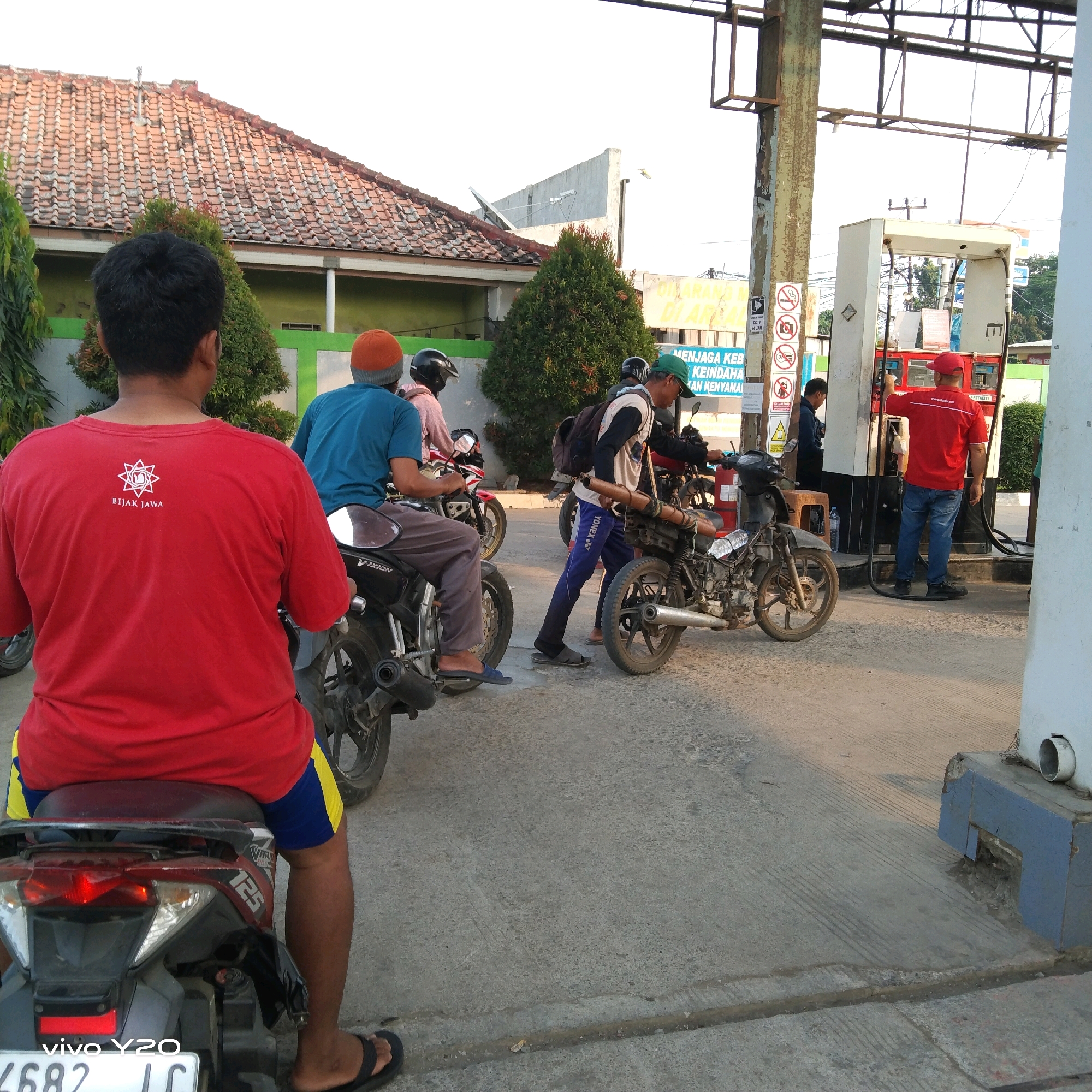 The image captures a scene at a gas station in an outdoor urban environment. Several people are present, primarily interacting with motorcycles and a gas pump.
In the foreground, a person wearing a red t-shirt with a white emblem and text that reads "BIJAK JAWA" is seated on a dark-colored motorcycle. They are facing away from the camera, and their legs are visible, wearing blue and yellow shorts and black flip-flops.
Behind the person in the red shirt, another individual in a blue t-shirt and gray pants is also seated on a motorcycle. They are wearing an orange beanie. Further back, a person in a pink shirt and a black helmet is on a motorcycle.
To the right, a person in a white long-sleeved shirt, blue pants, and a green cap is walking, carrying a backpack and a long, cylindrical object. They are near a gas pump. Another person, wearing a red cap and a red shirt with "PERTAMINA" visible, is operating the gas pump, with a hose connected to a vehicle, likely a motorcycle. This person is wearing blue jeans and black shoes.
The gas station features a red and white gas pump. Signs are visible on a pole near the pump, including one with no smoking symbols and another with text in Indonesian. The environment appears to be a paved area, with some greenery and a building with a tiled roof in the background. The lighting suggests it is daytime, possibly late afternoon, given the shadows. The license plate of the motorcycle in the foreground is partially visible and reads "4682 LC". The phone brand "vivo Y20" is also visible on the license plate.