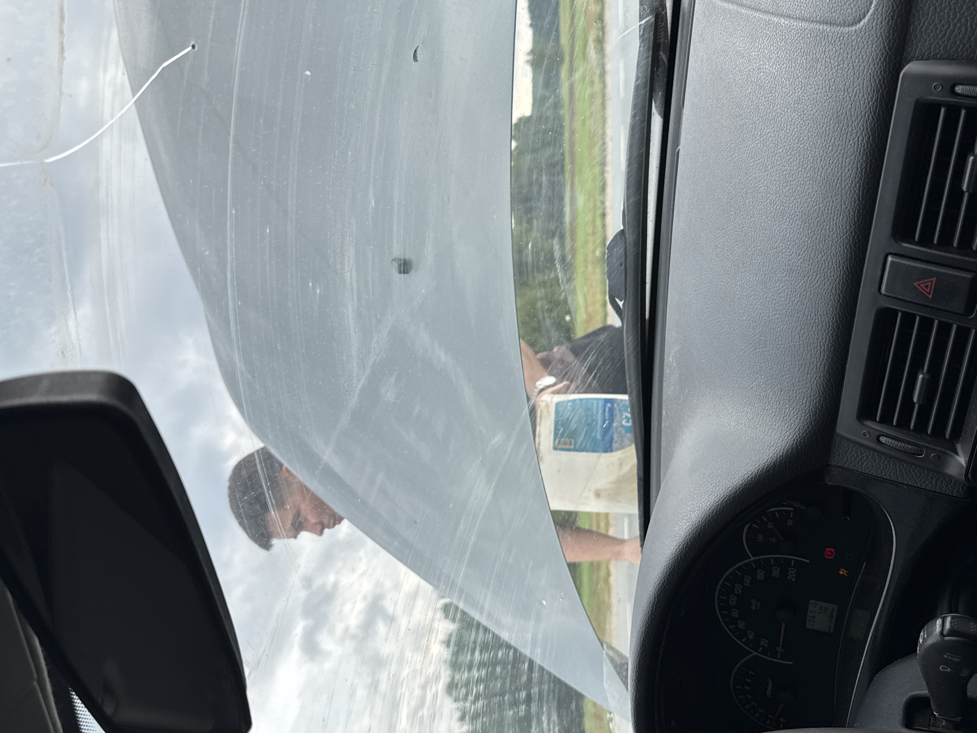 The image, taken from inside a car, shows a young man standing outside with the hood of the car open. The man is in profile, looking towards the car's engine. He is wearing a light-colored shirt and holding a light-colored bag. The car's dashboard, including the speedometer, is visible in the foreground. The scene appears to be on the side of a road with trees and fields in the background. The sky is overcast. The location is identified as Shumiha, Russia.