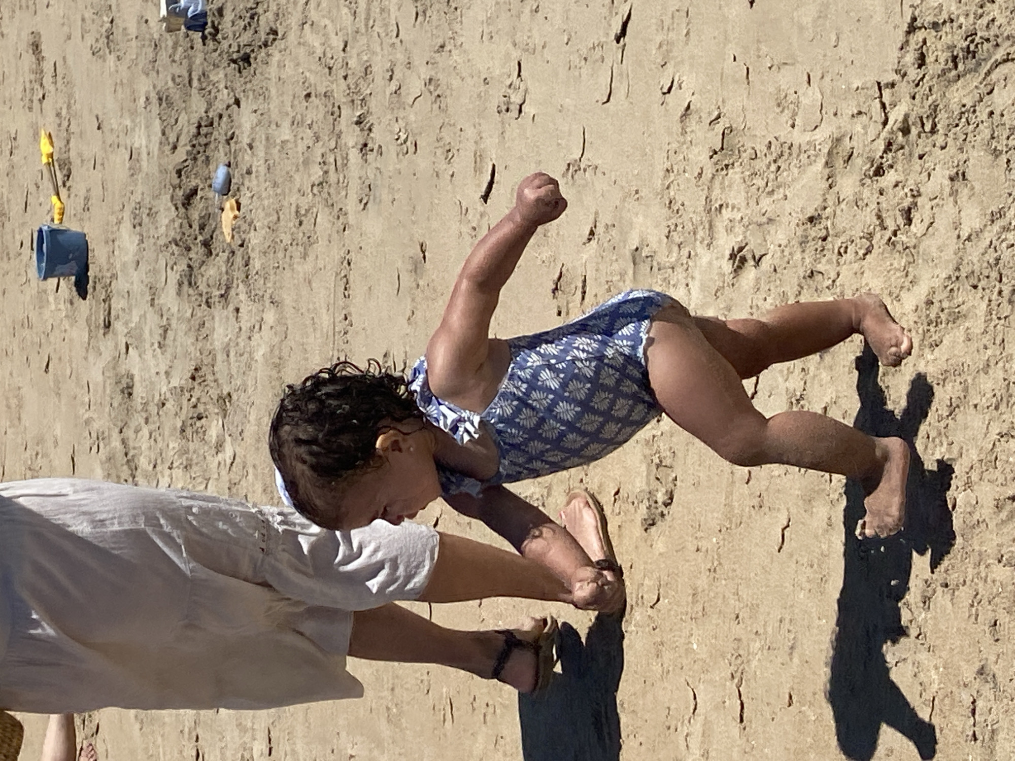The image shows a young child in a blue and white patterned swimsuit on a sandy beach, likely in Conil de la Frontera, Spain. The child is in a dynamic pose, leaning forward with one arm raised and clenched into a fist, appearing to be mid-movement or play. Their legs are bent, with one foot visible in the sand. Another person, partially visible from the waist down, is wearing a white garment and sandals, and is reaching out towards the child, possibly helping them or playing with them.
The setting is a beach with the sand showing textures and indentations, suggesting it might be wet or recently disturbed. There are some small beach toys scattered on the sand in the background, including a blue bucket and a yellow shovel. The sunlight casts a strong shadow of the child and the other person on the sand, indicating it is likely a sunny day with the sun relatively high in the sky, suggesting midday. The overall mood appears to be one of active, playful engagement, and possibly a parent or guardian interacting with a child on a beach outing. No visible text is present in the image.